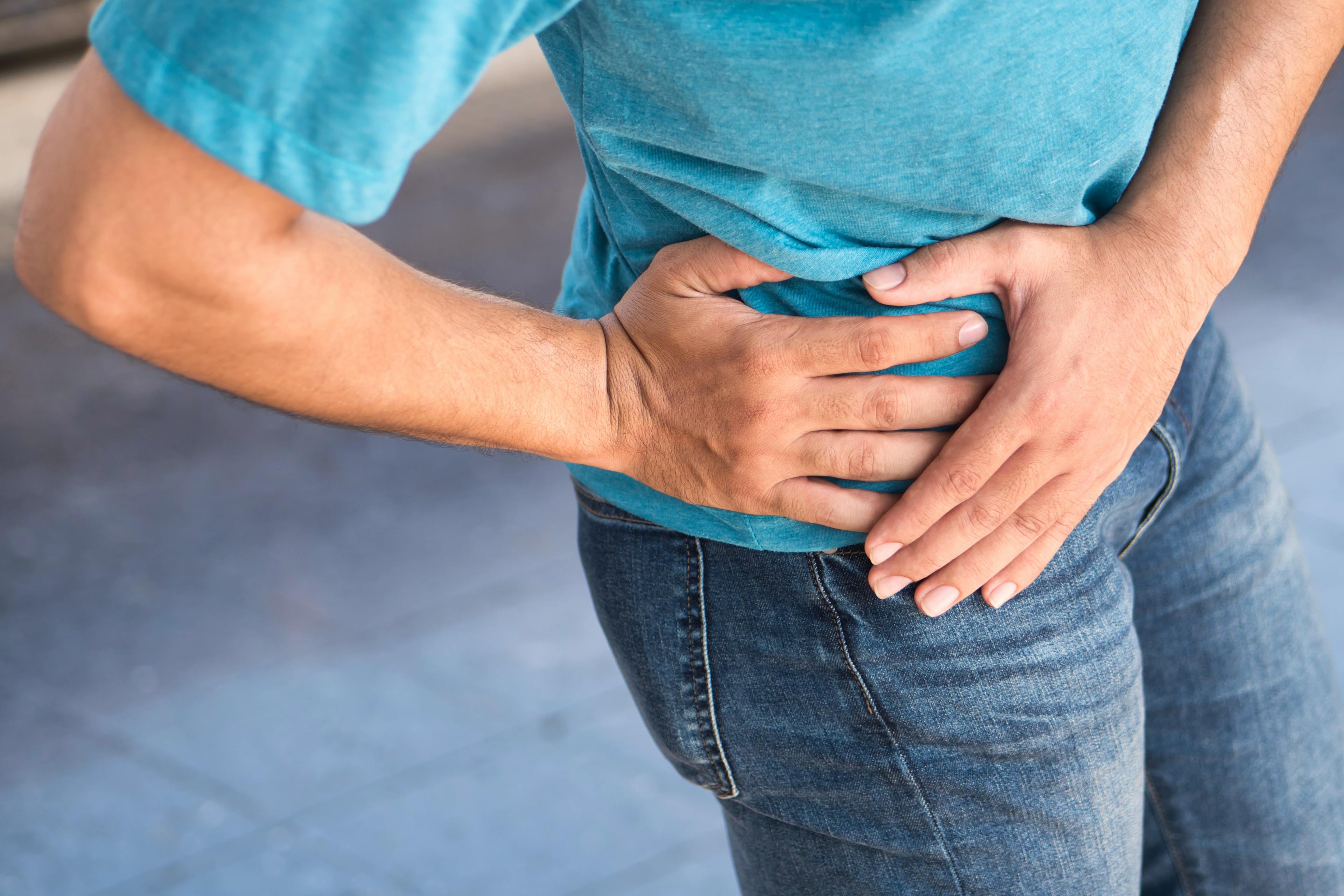 Person clutching their right hip in pain, wearing a blue shirt and jeans, possibly experiencing discomfort or injury in the hip or lower abdomen.