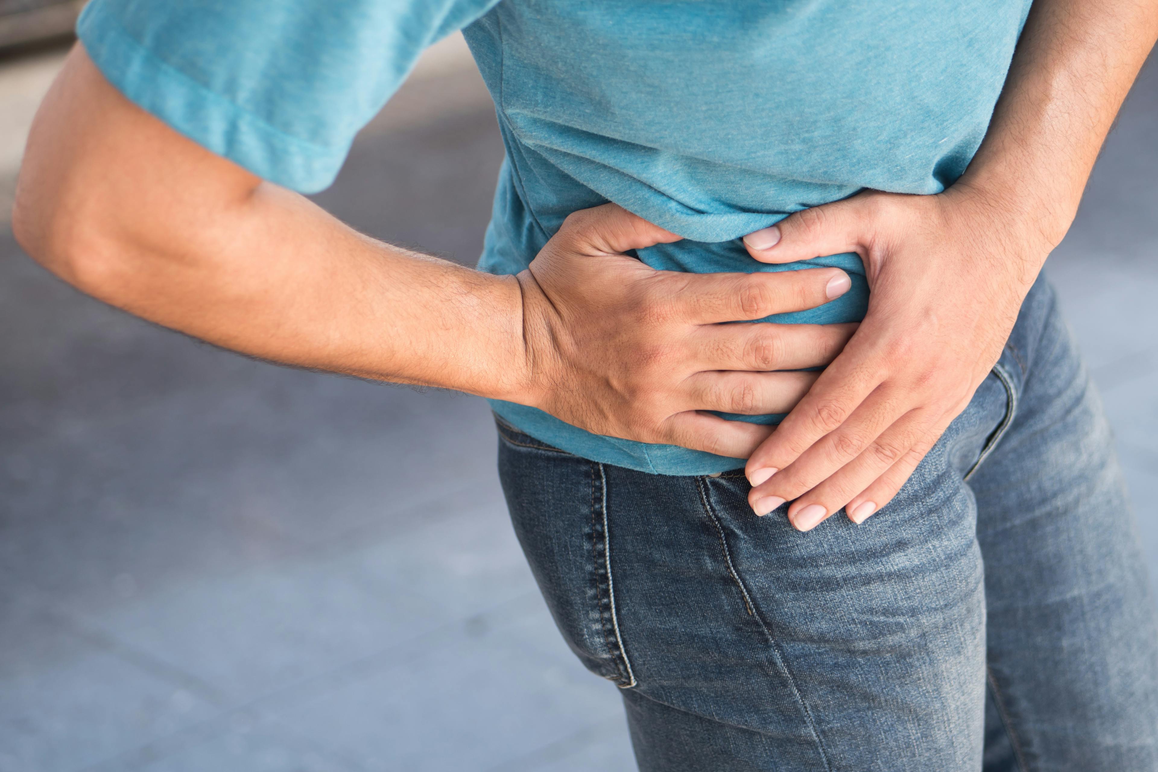 Person clutching their right hip in pain, wearing a blue shirt and jeans, possibly experiencing discomfort or injury in the hip or lower abdomen.
