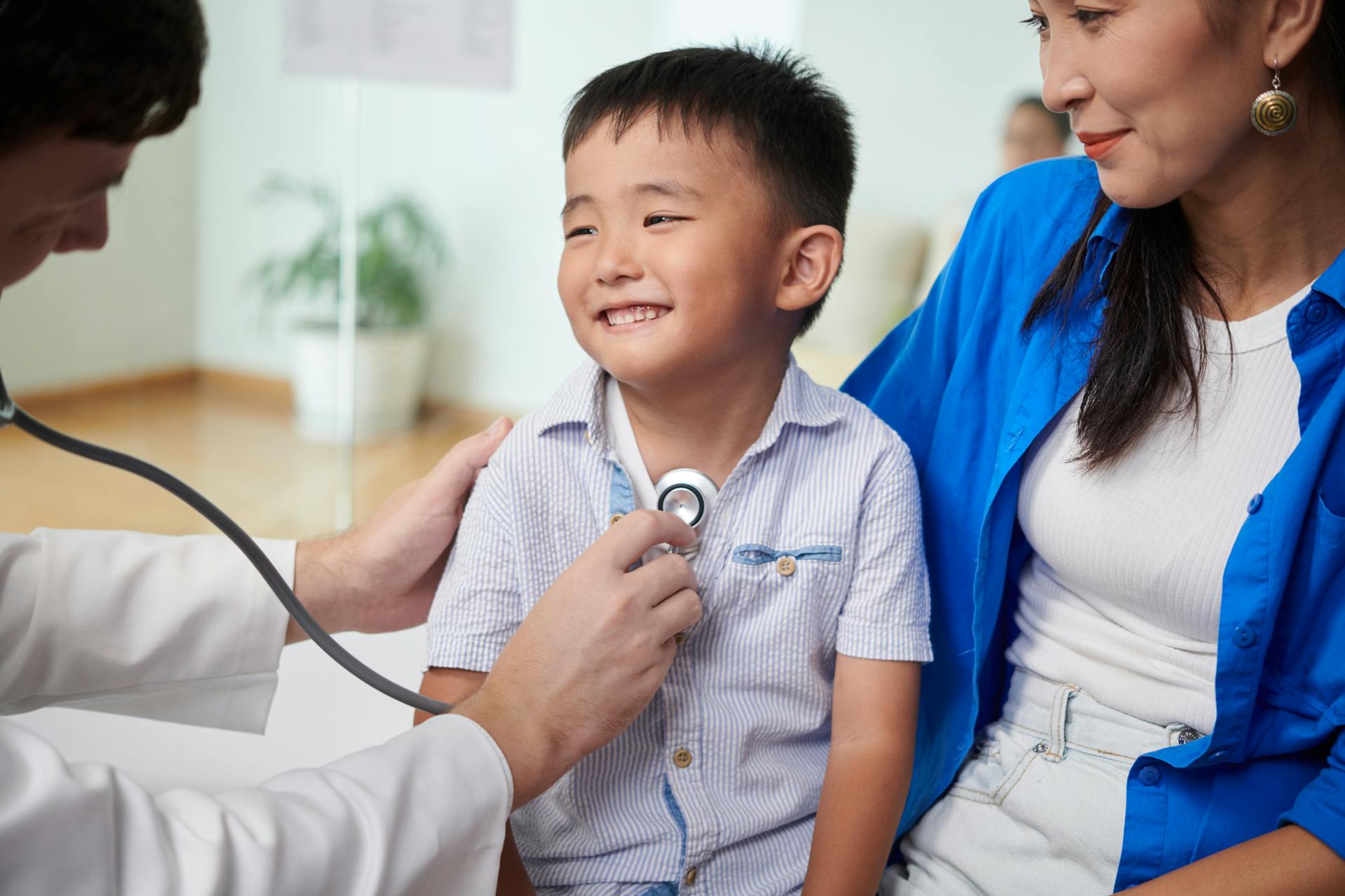 A smiling child in his mom's lap with a doctor using a stethoscope on him.