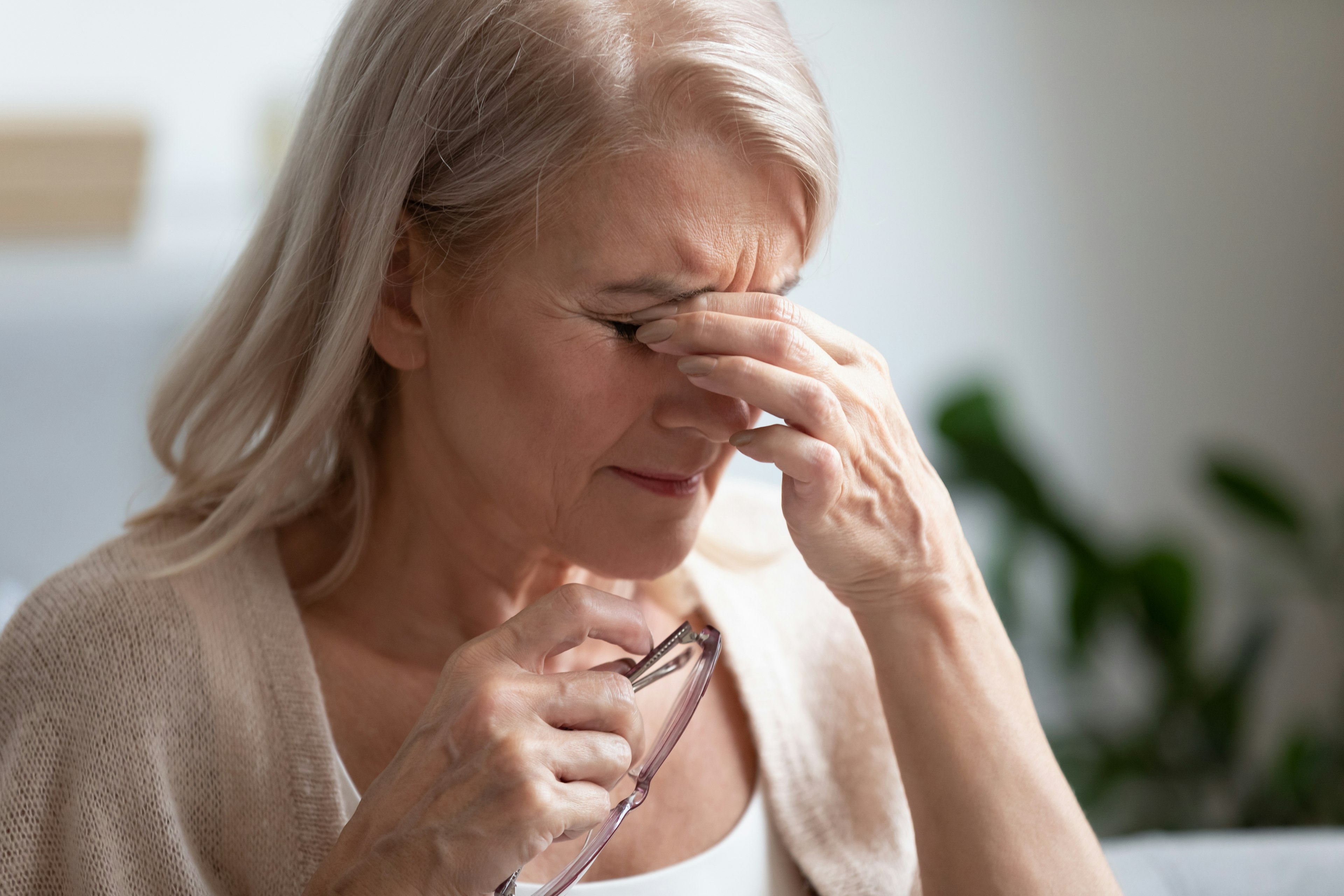 Older woman with light gray hair holding her eyeglasses in one hand and pressing her fingers to the bridge of her nose, appearing to experience eye discomfort.