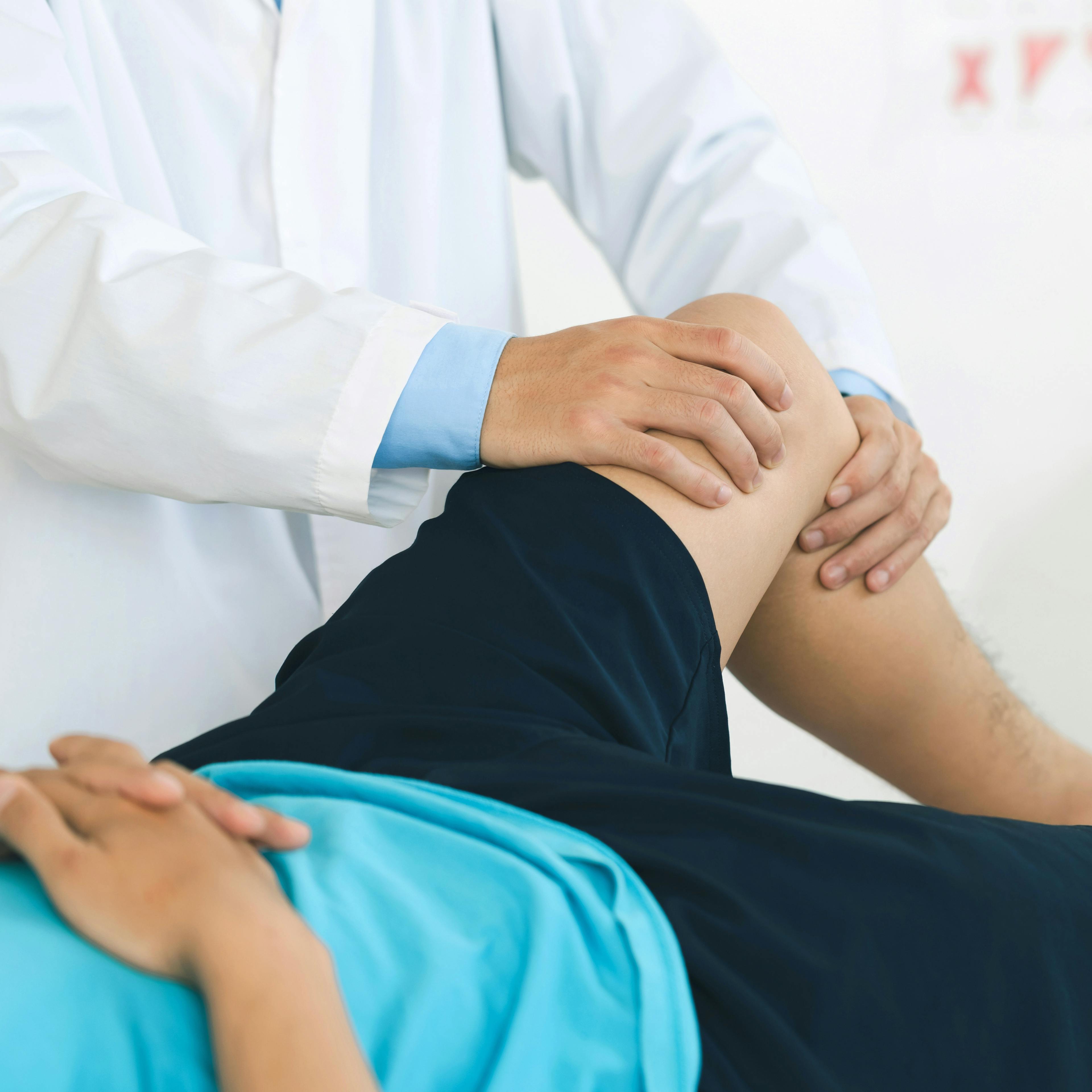 Healthcare provider examining a young patient's knee during a physical exam, with the patient lying on an exam table and wearing athletic shorts and a blue shirt.