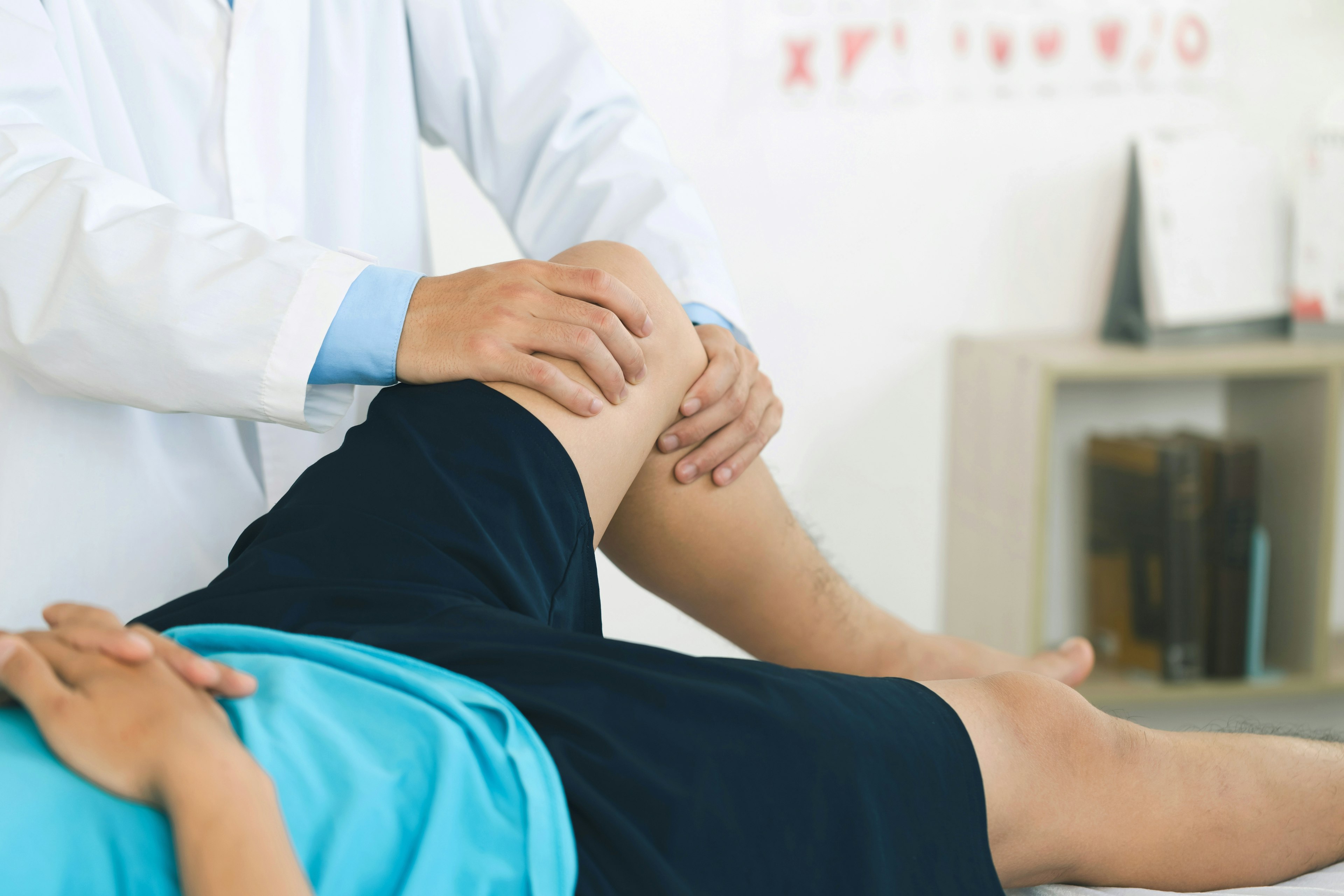 Healthcare provider examining a young patient's knee during a physical exam, with the patient lying on an exam table and wearing athletic shorts and a blue shirt.