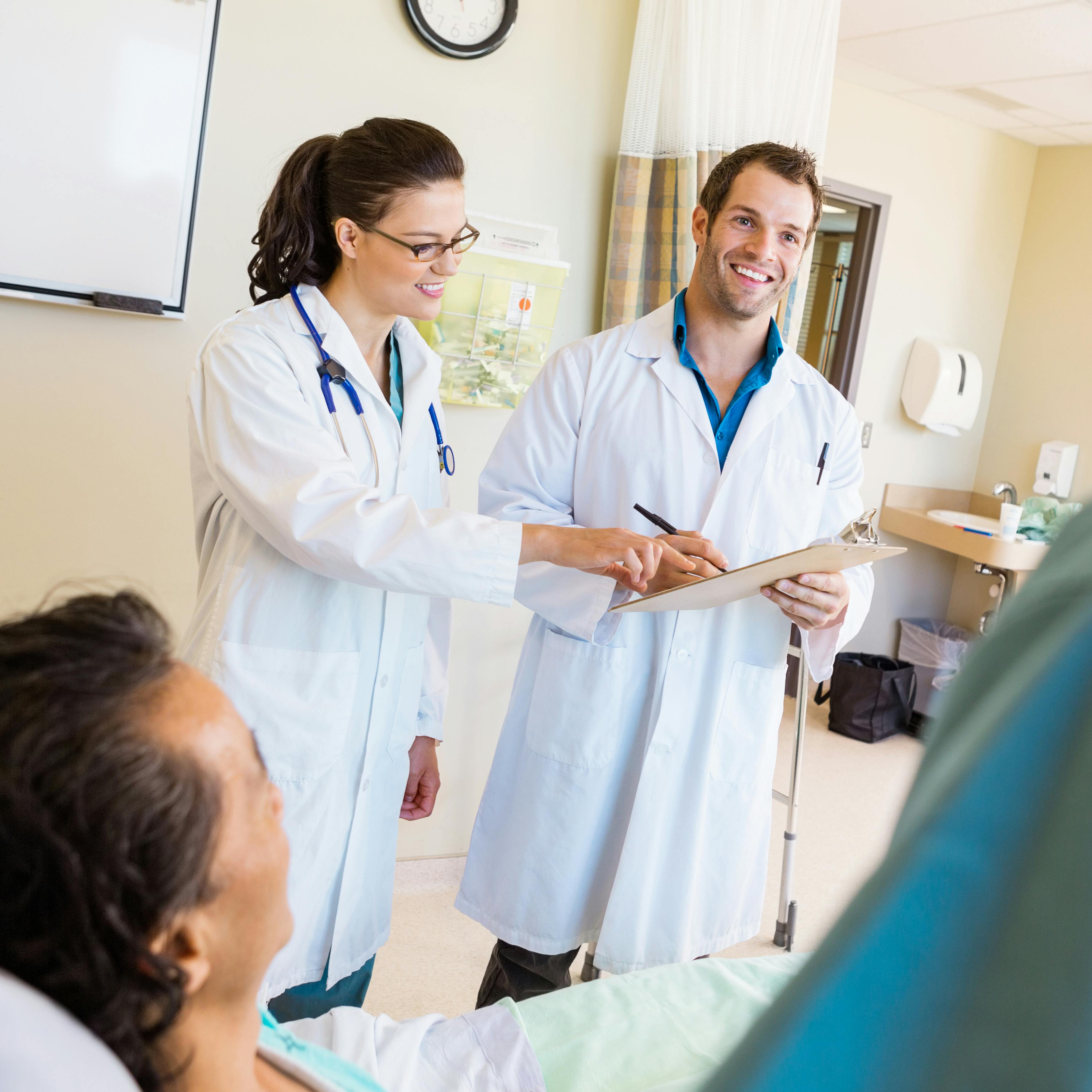Medical residents are assisting in a bedside patient's care while speaking to their preceptor.