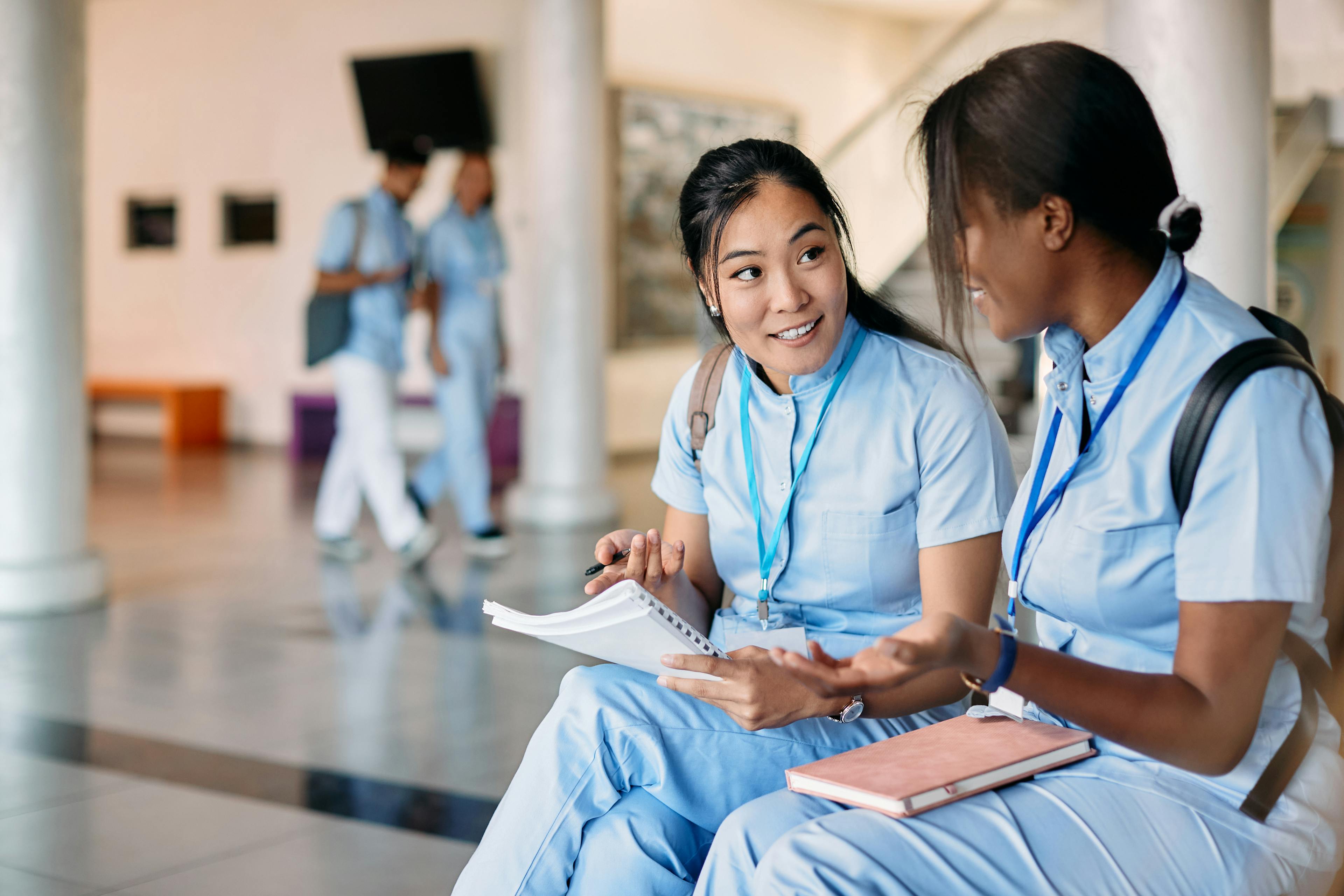 A nursing student is sitting next to her peer, discussing the educational content in her notebook. 