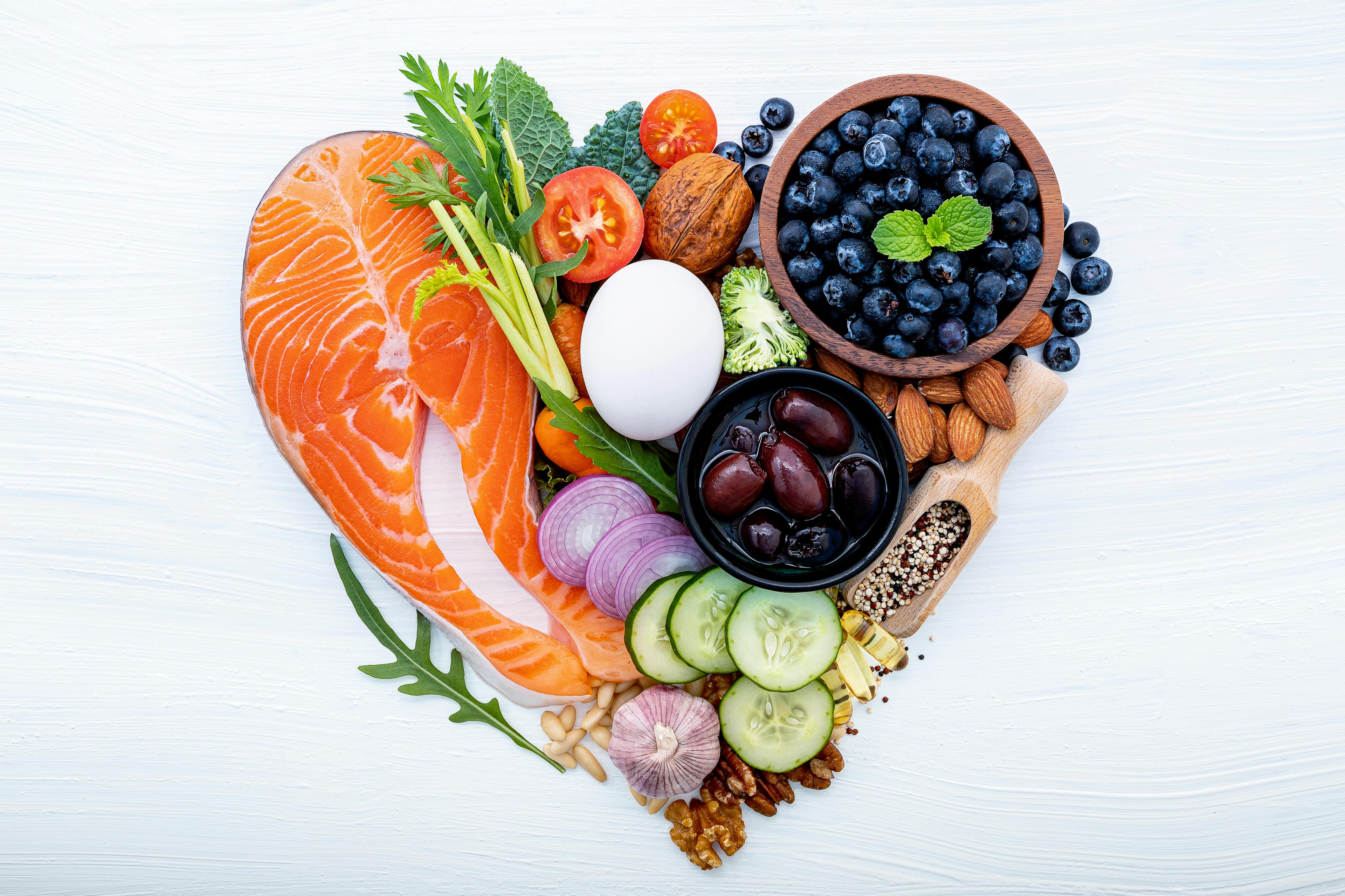 A heart-shaped arrangement of healthy foods on a white background, including a salmon fillet, blueberries, an egg, black olives, cherry tomatoes, red onion, cucumber slices, garlic, nuts (walnuts, almonds), seeds, celery, leafy greens, and various colorful vegetables—symbolizing heart-healthy nutrition.