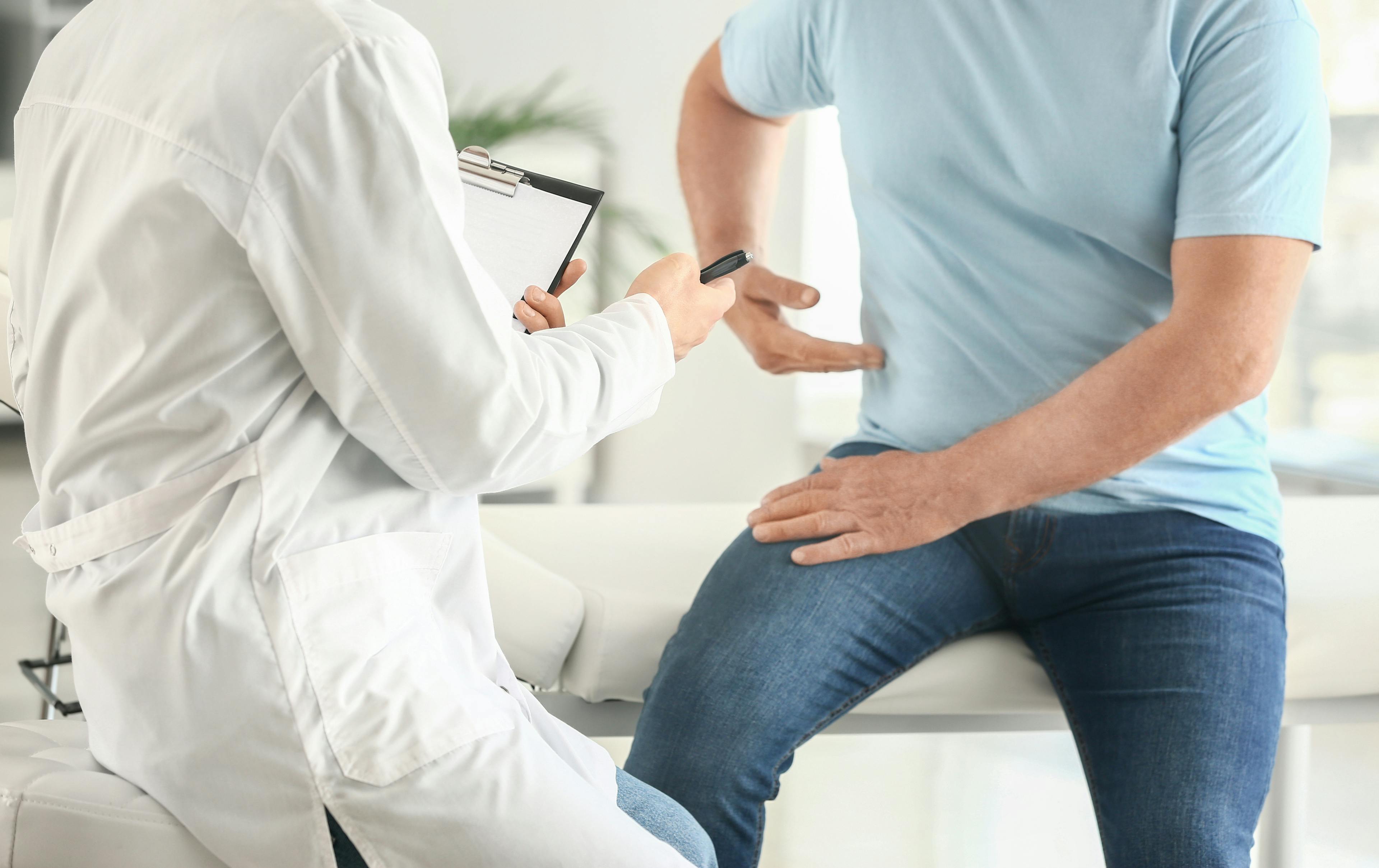 A healthcare provider in a white coat holding a chart opposite a patient gesturing to an area in his midsection. 
