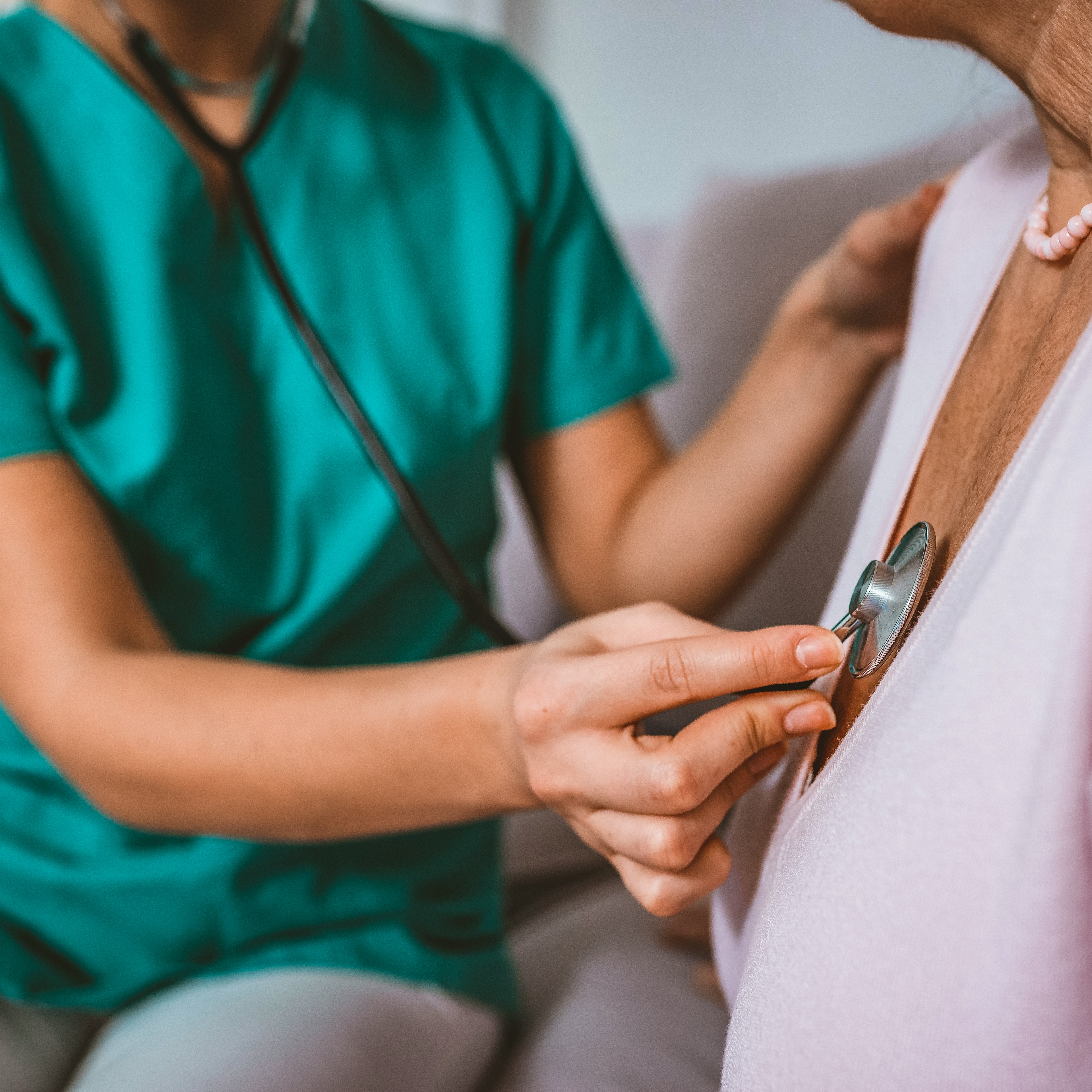 An elderly woman is having her heartbeat checked with a stethoscope by a person in teal scrubs.