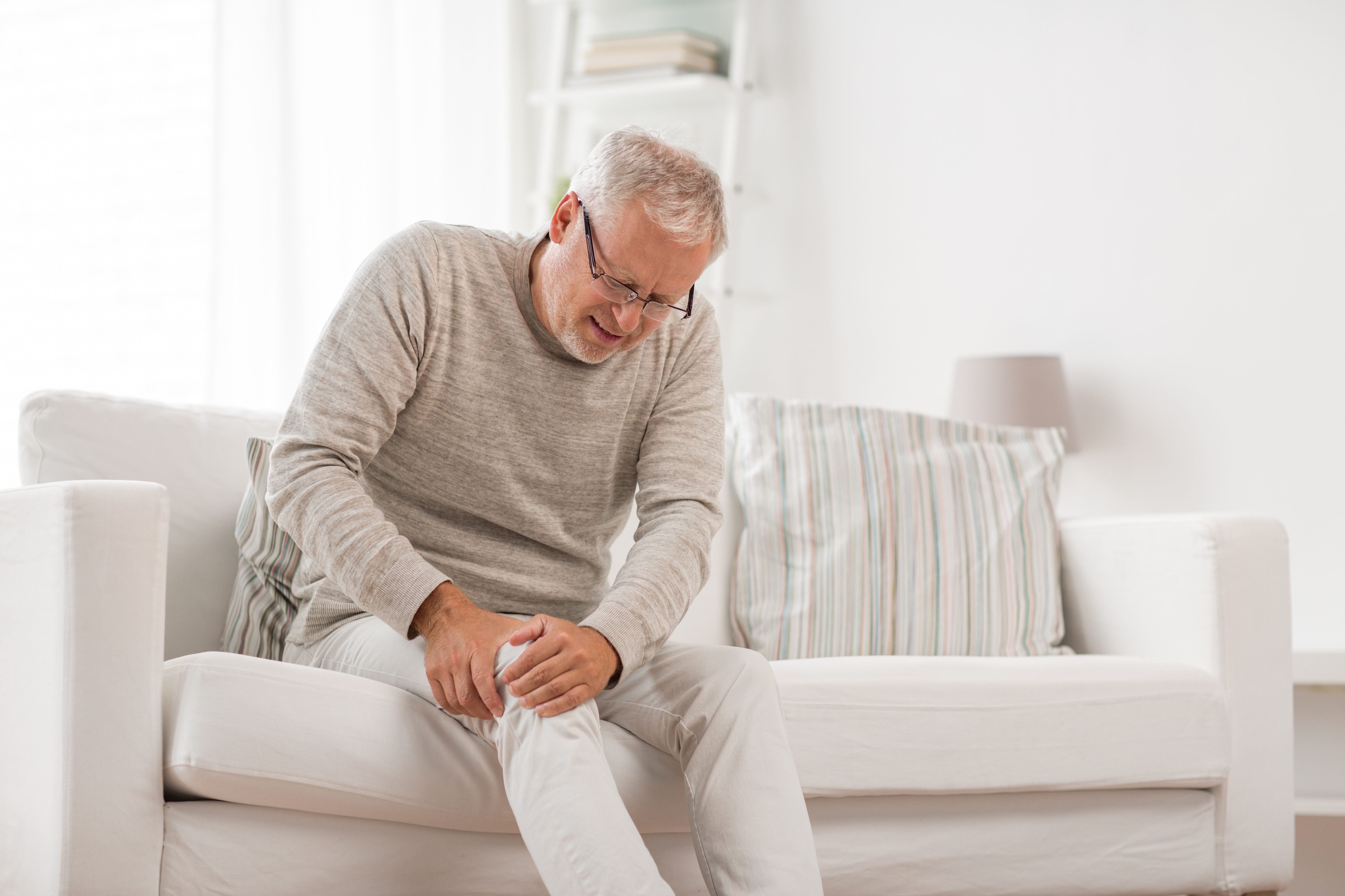 An elderly man is sitting on a couch, holding his knee due to joint pain. 