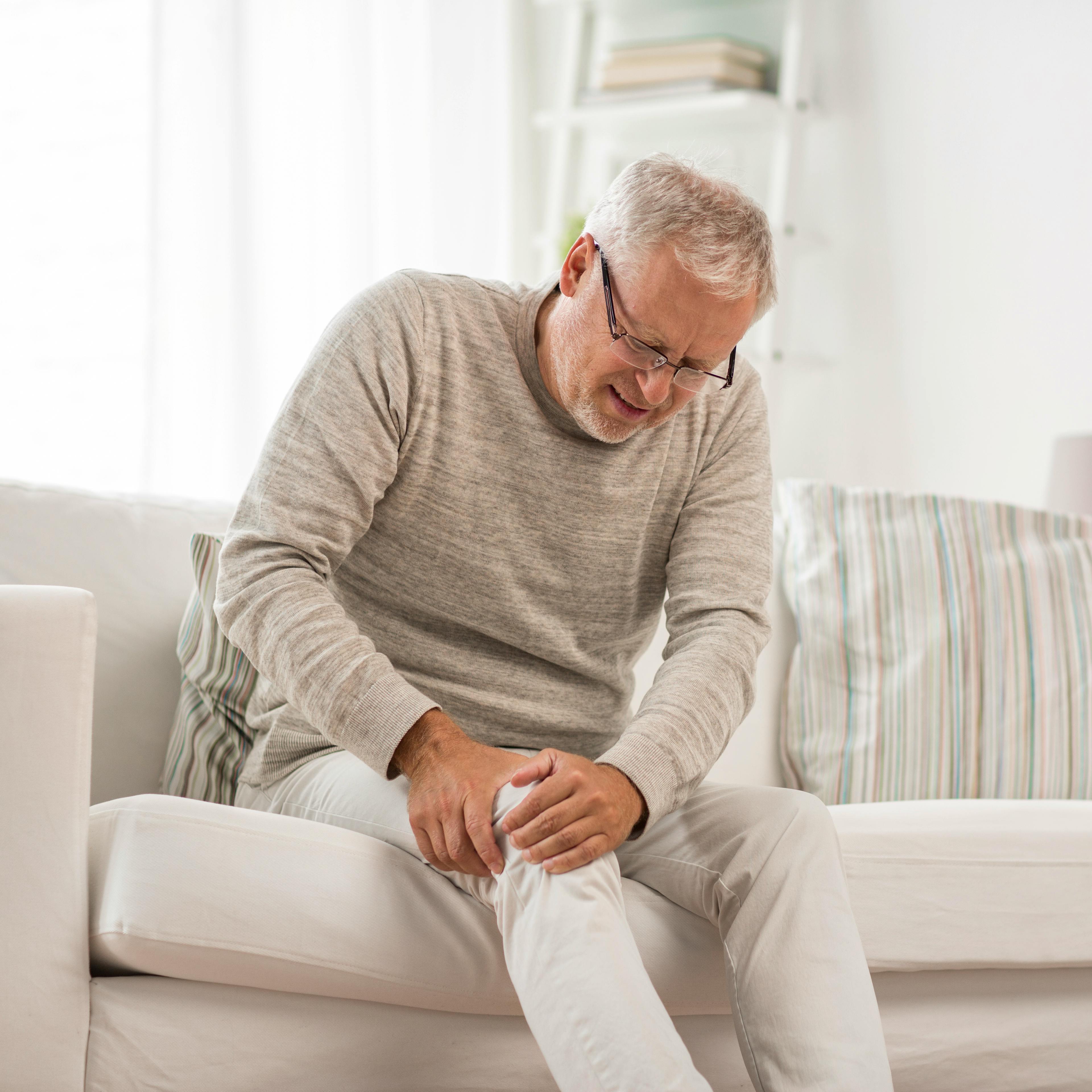 An elderly man is sitting on a couch, holding his knee due to joint pain.