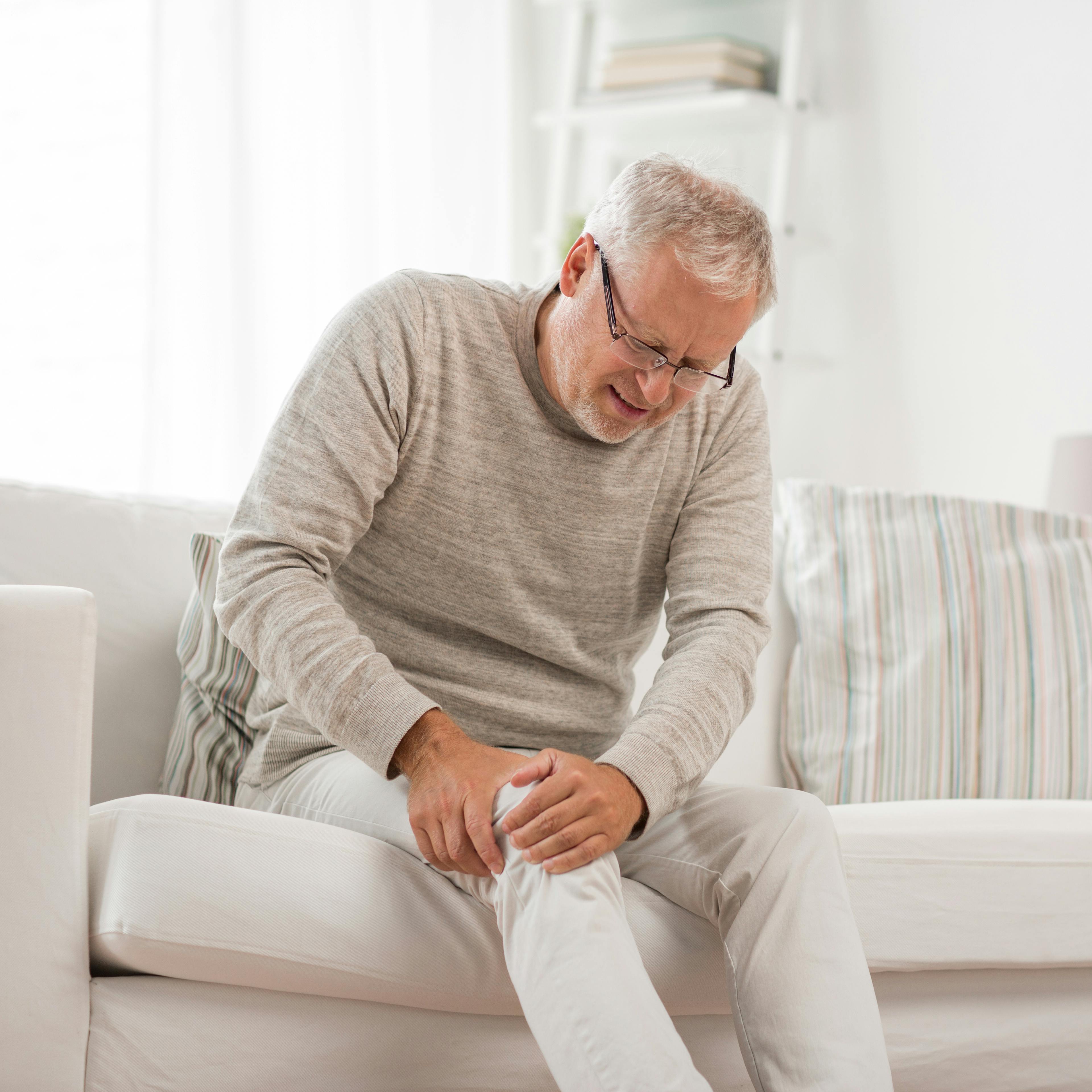 An elderly man is sitting on a couch, holding his knee due to joint pain.