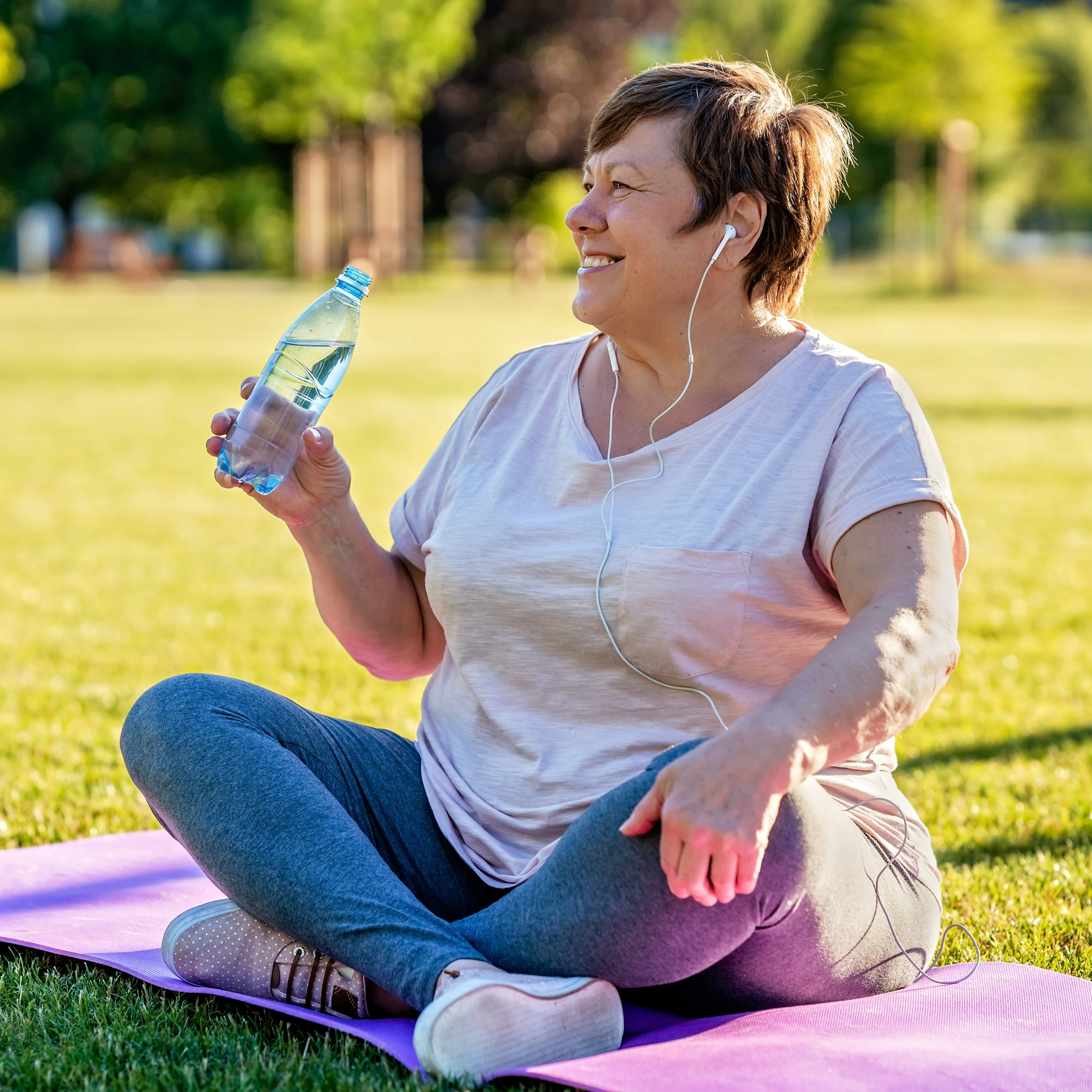 A woman is sitting in a park on a yoga mat while hydrating. 