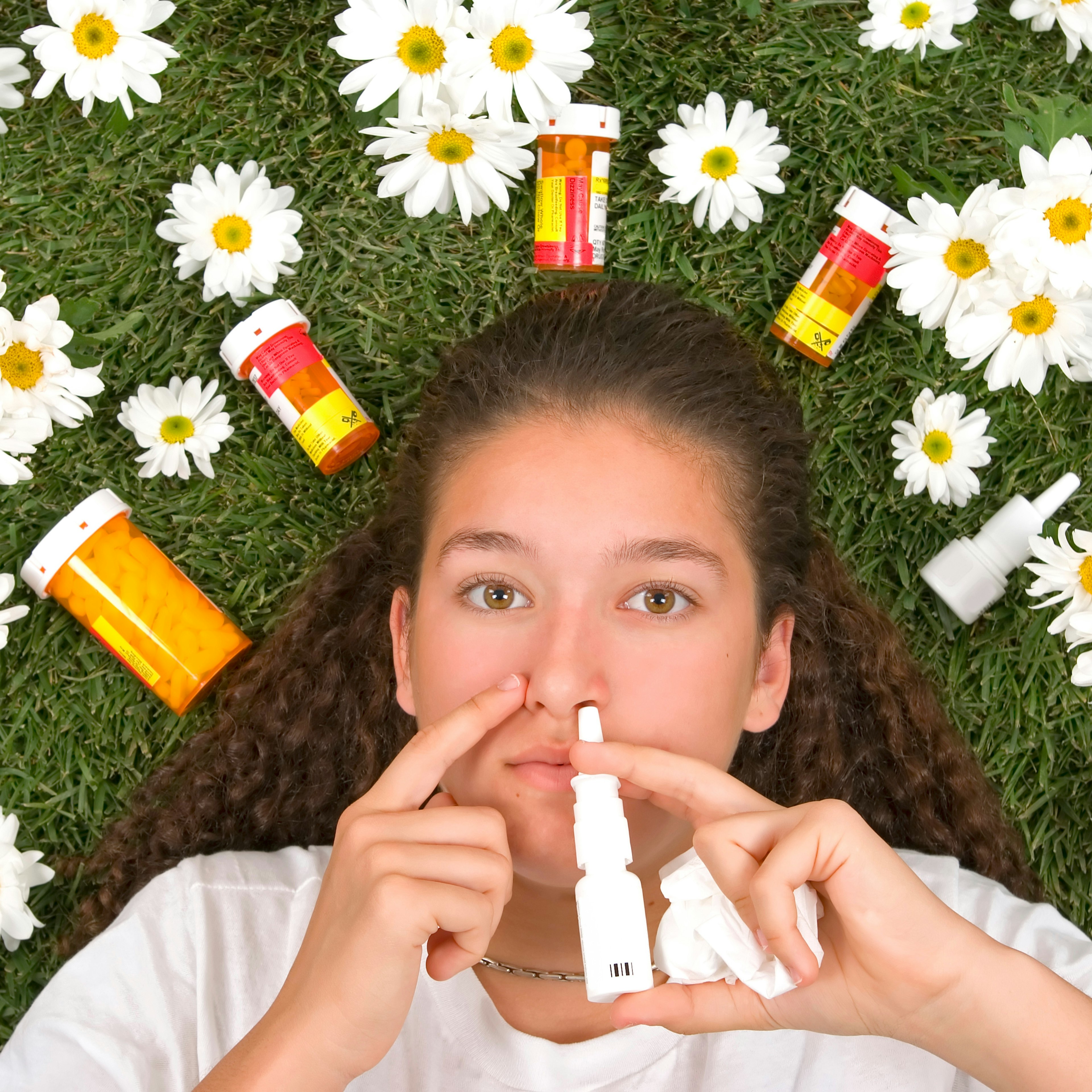A girl is laying in grass surrounded by flowers with various allergy medications. 