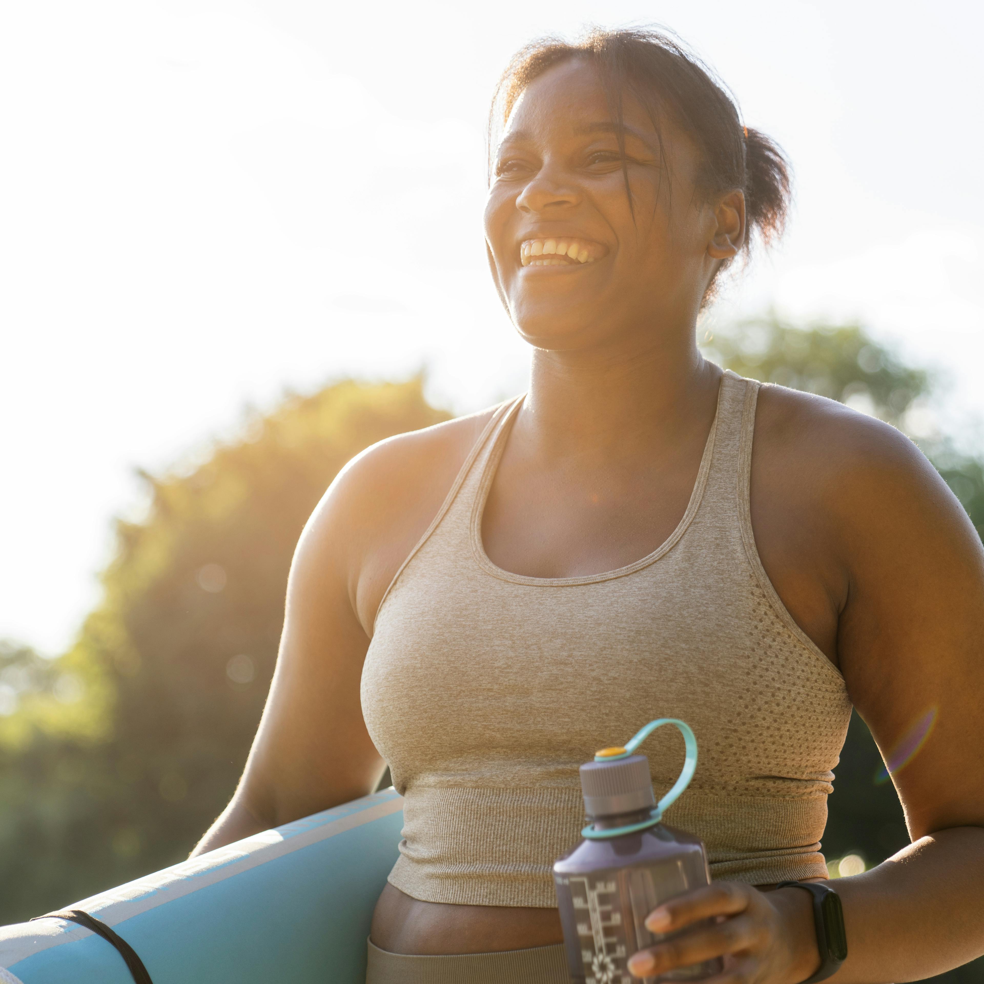 A woman is happily outdoors holding a yoga mat and water bottle. 