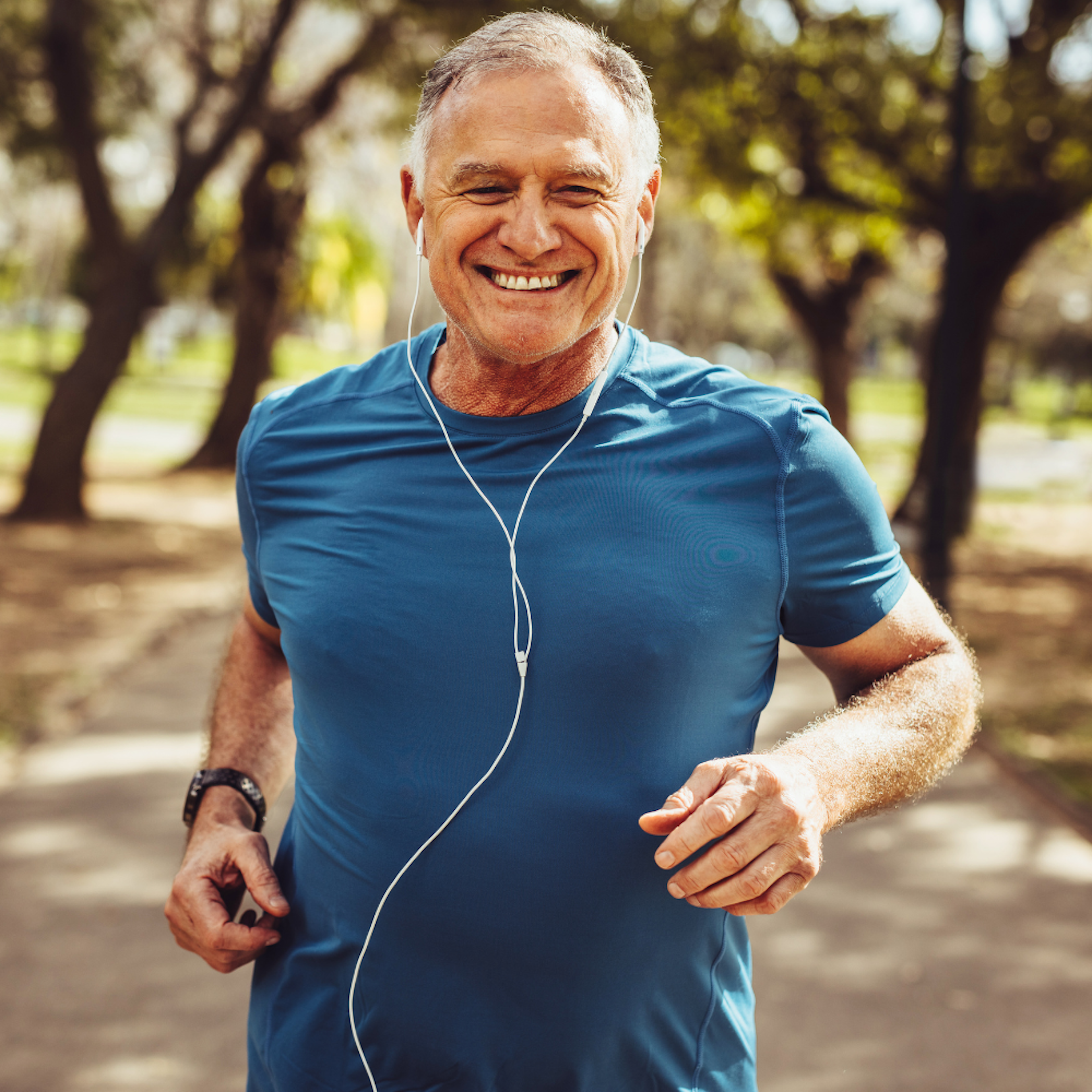An older man is running through a park with a smile on his face, showing a healthy lifestyle.