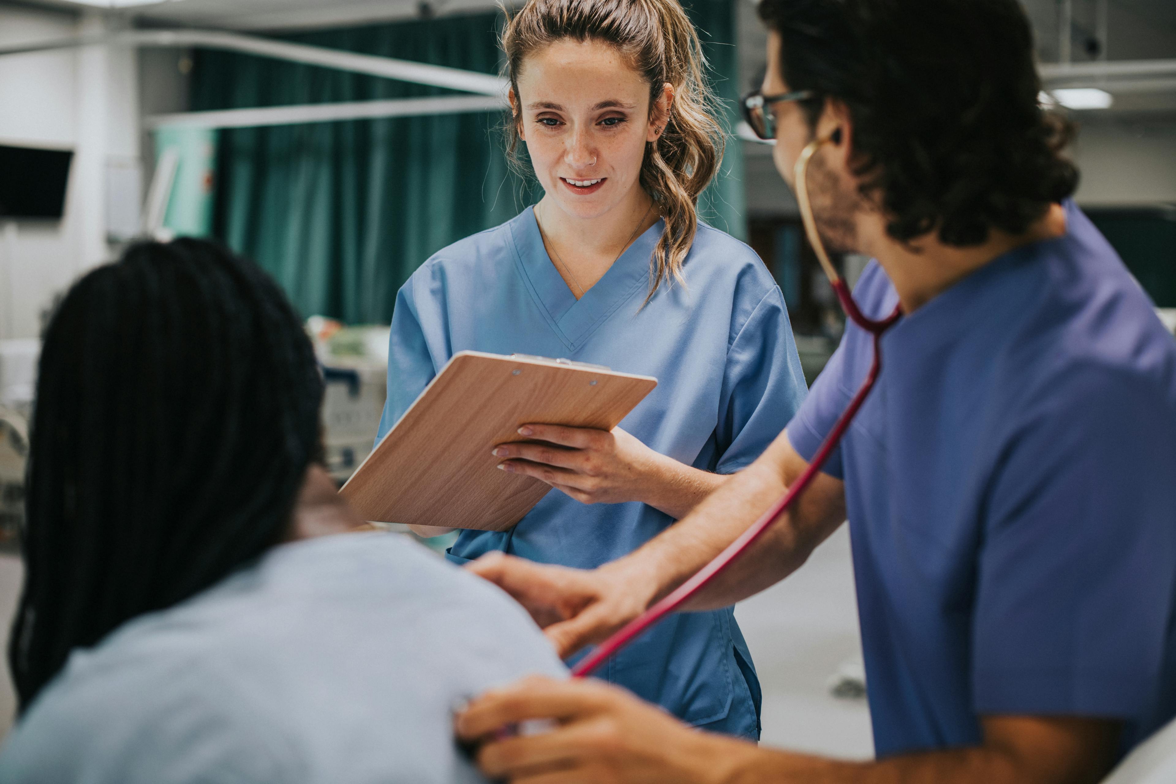 A nursing student is taking notes on a clipboard while observing her supervisor with a patient.