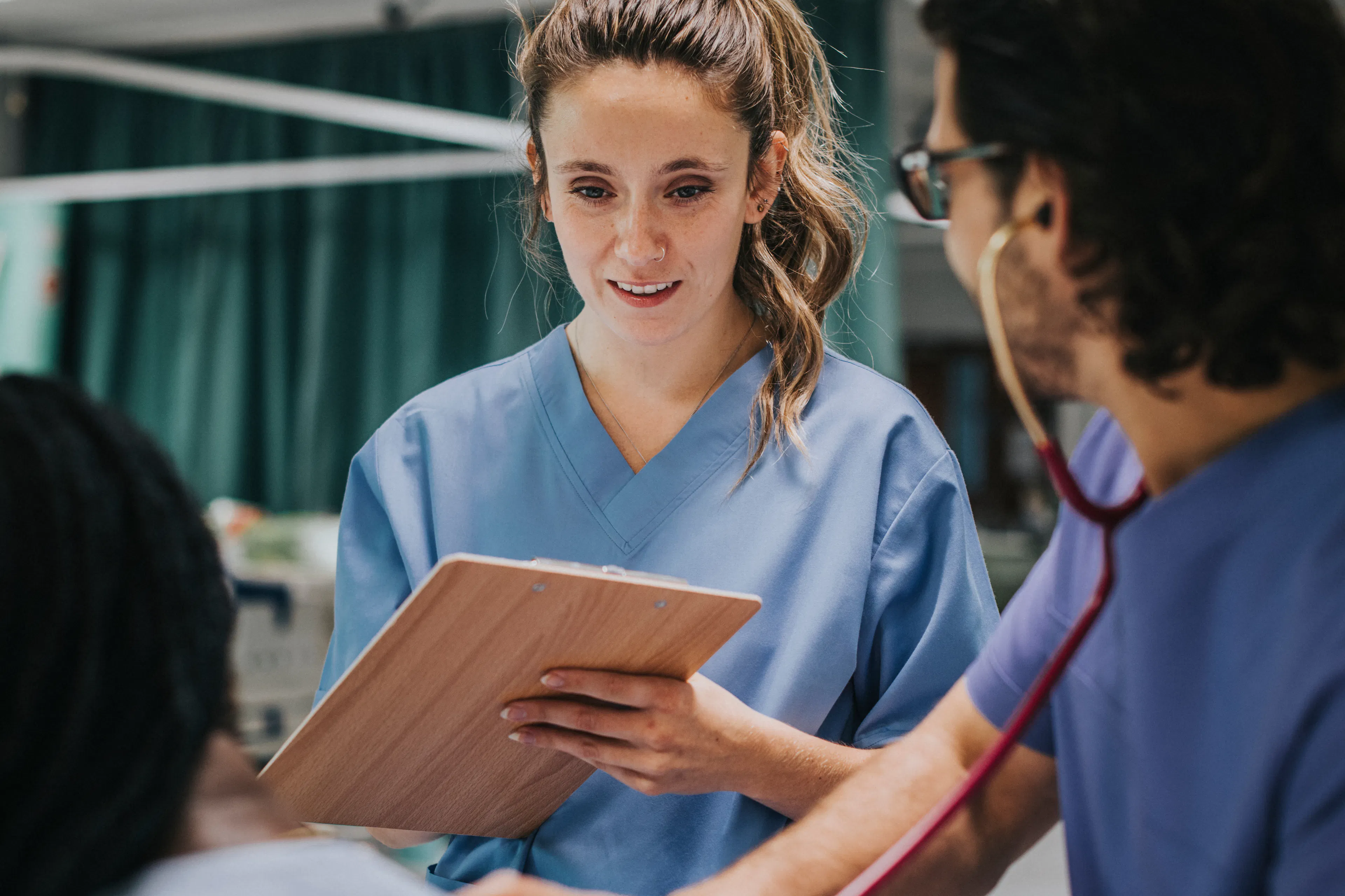 A nursing student is taking notes on a clipboard while observing her supervisor with a patient.