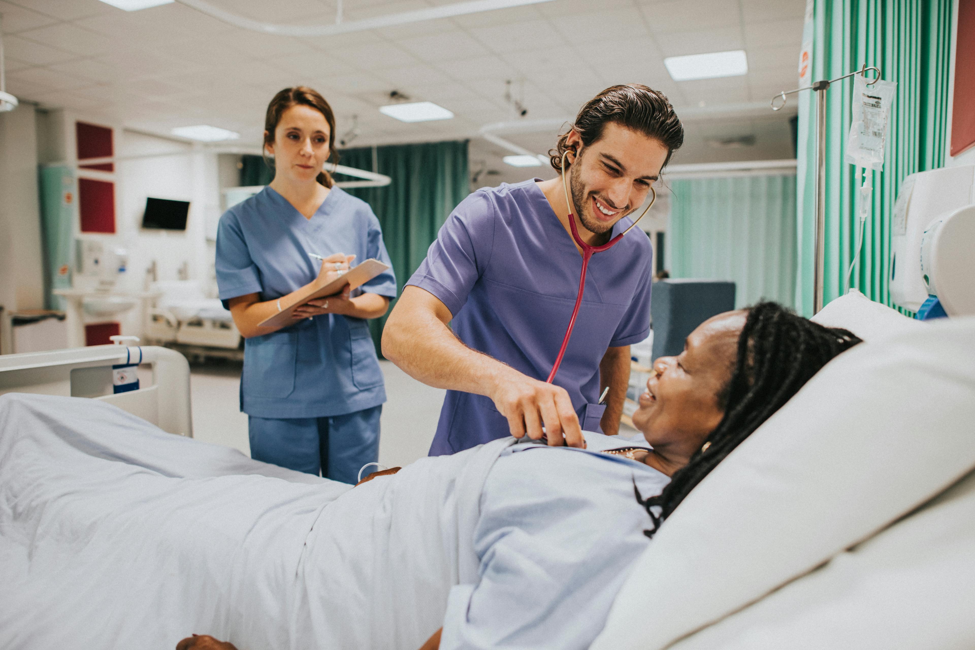 A male nursing student is using his stethoscope to check a patient while a female student takes notes.
