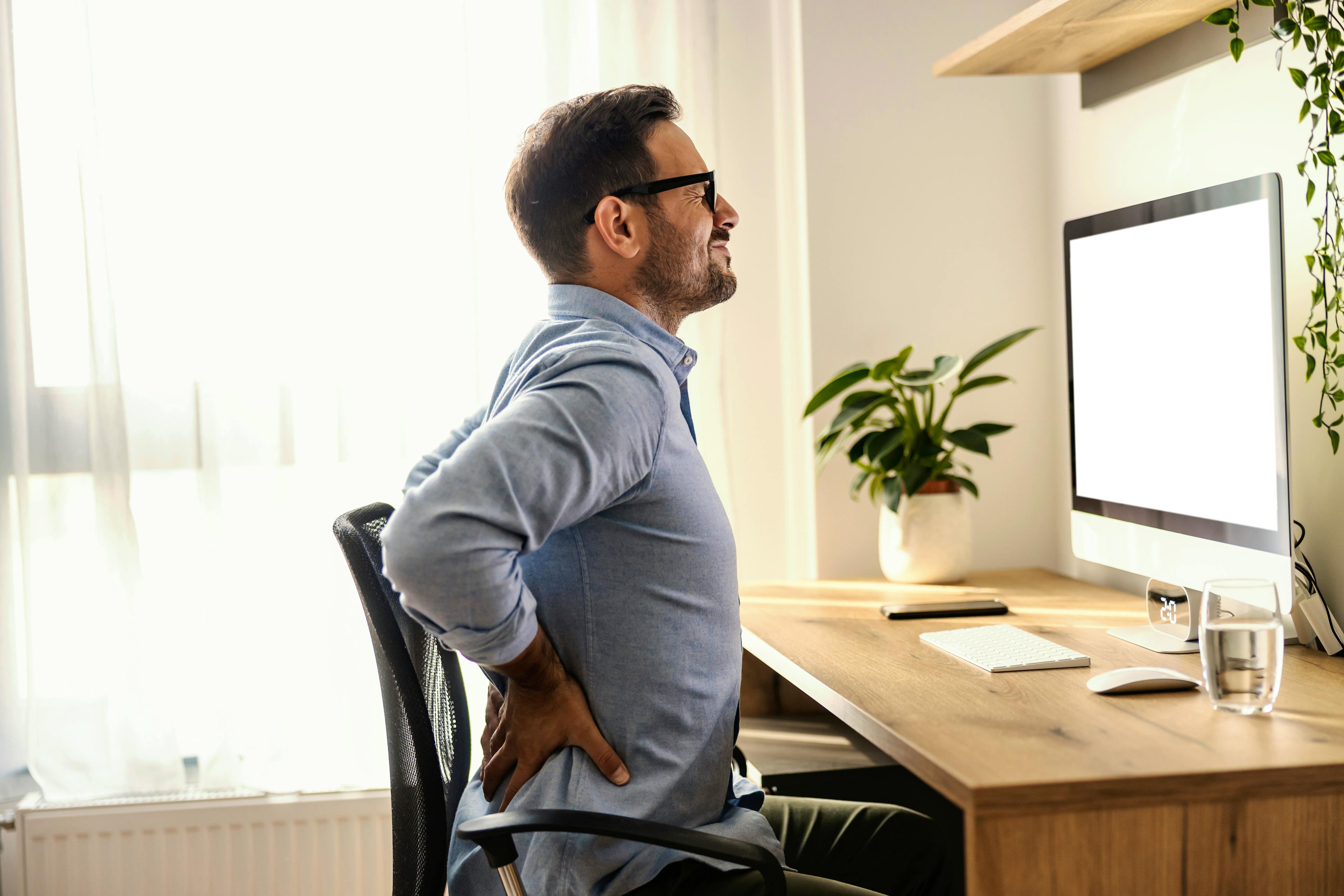 A man is experiencing lower back pain at his desk in what looks like his home office.