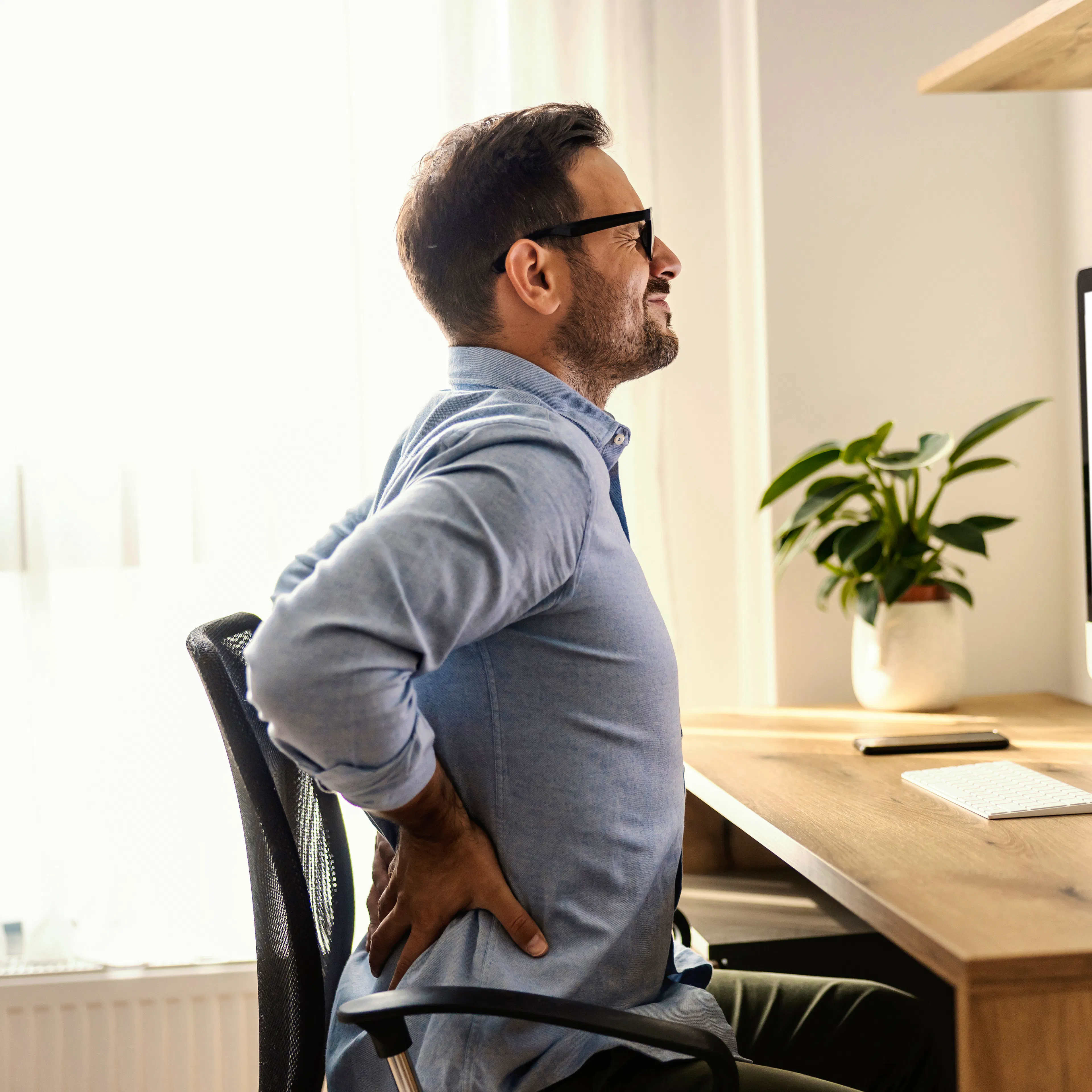 A man is experiencing lower back pain at his desk in what looks like his home office.