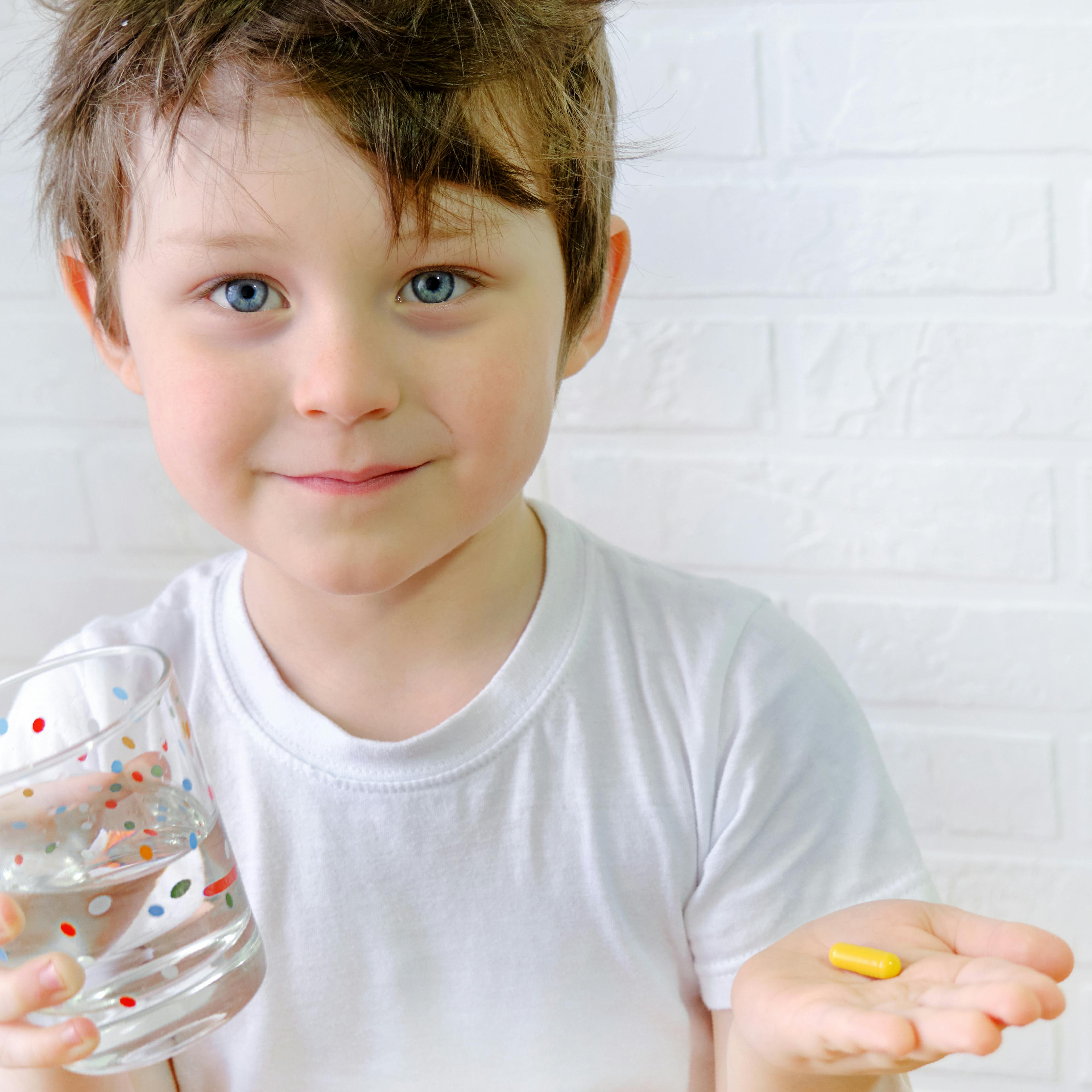 A child smiling holding a glass of water and his pill medication. 