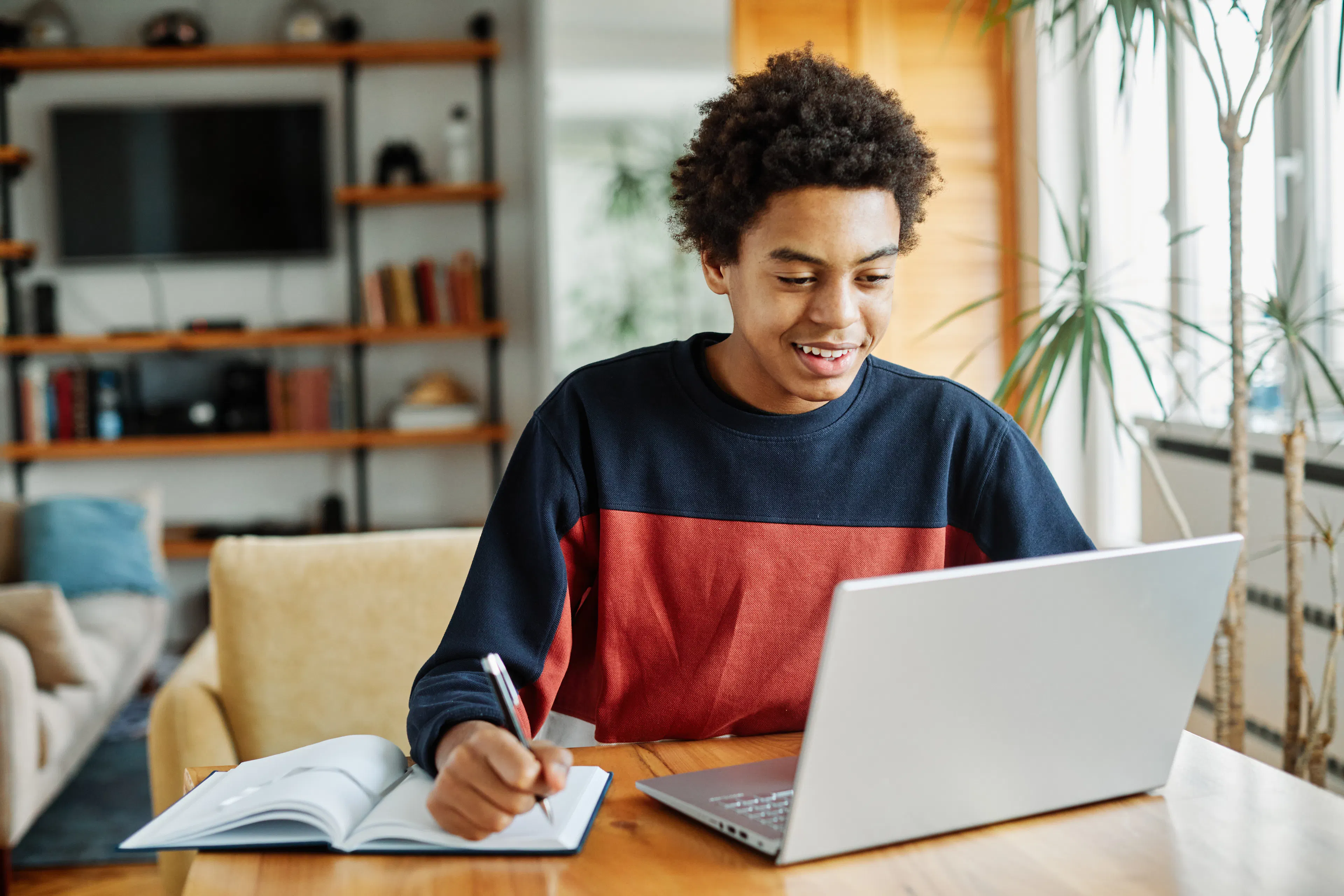 An adolescent teen is happily studying, writing notes while on his laptop.