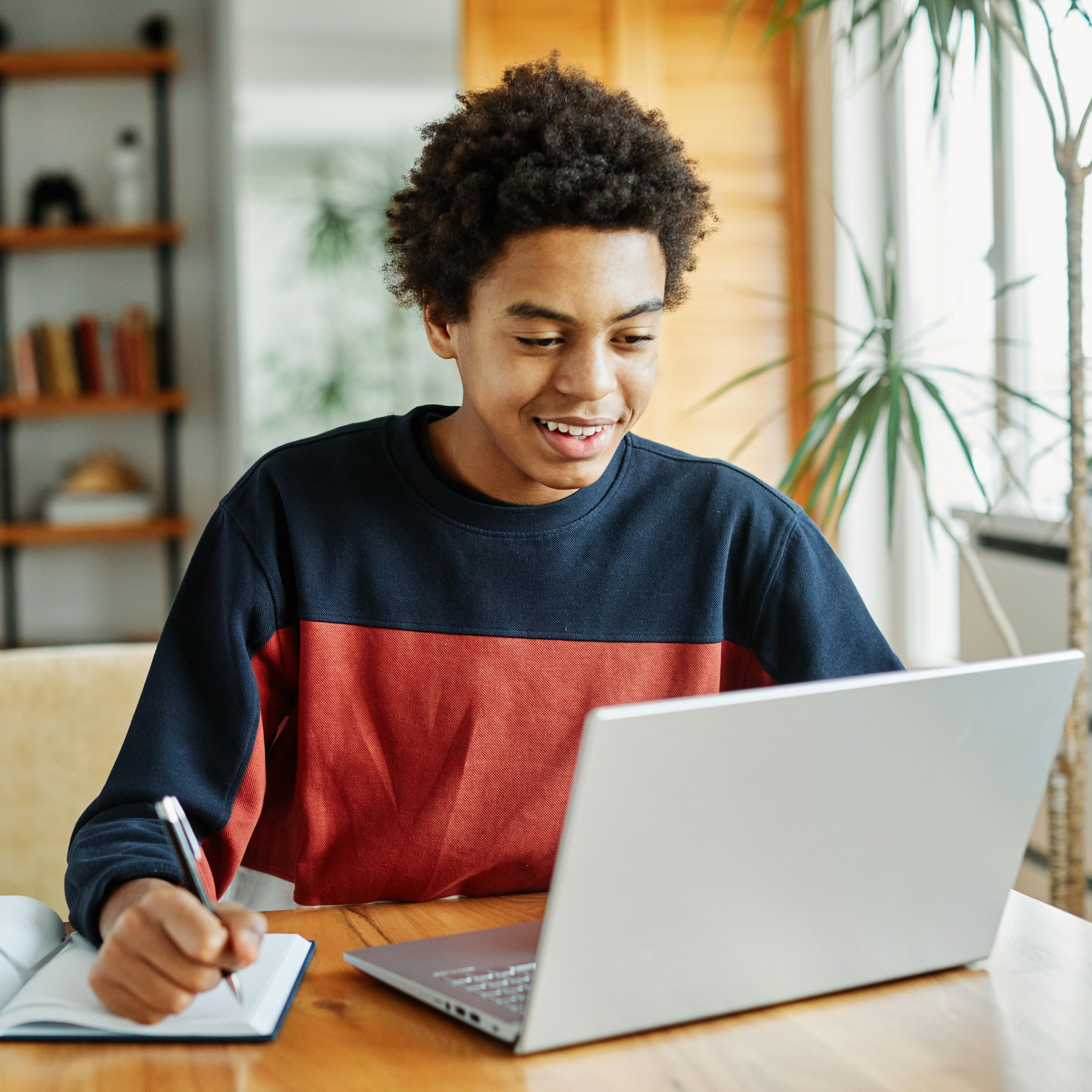 An adolescent teen is happily studying, writing notes while on his laptop.