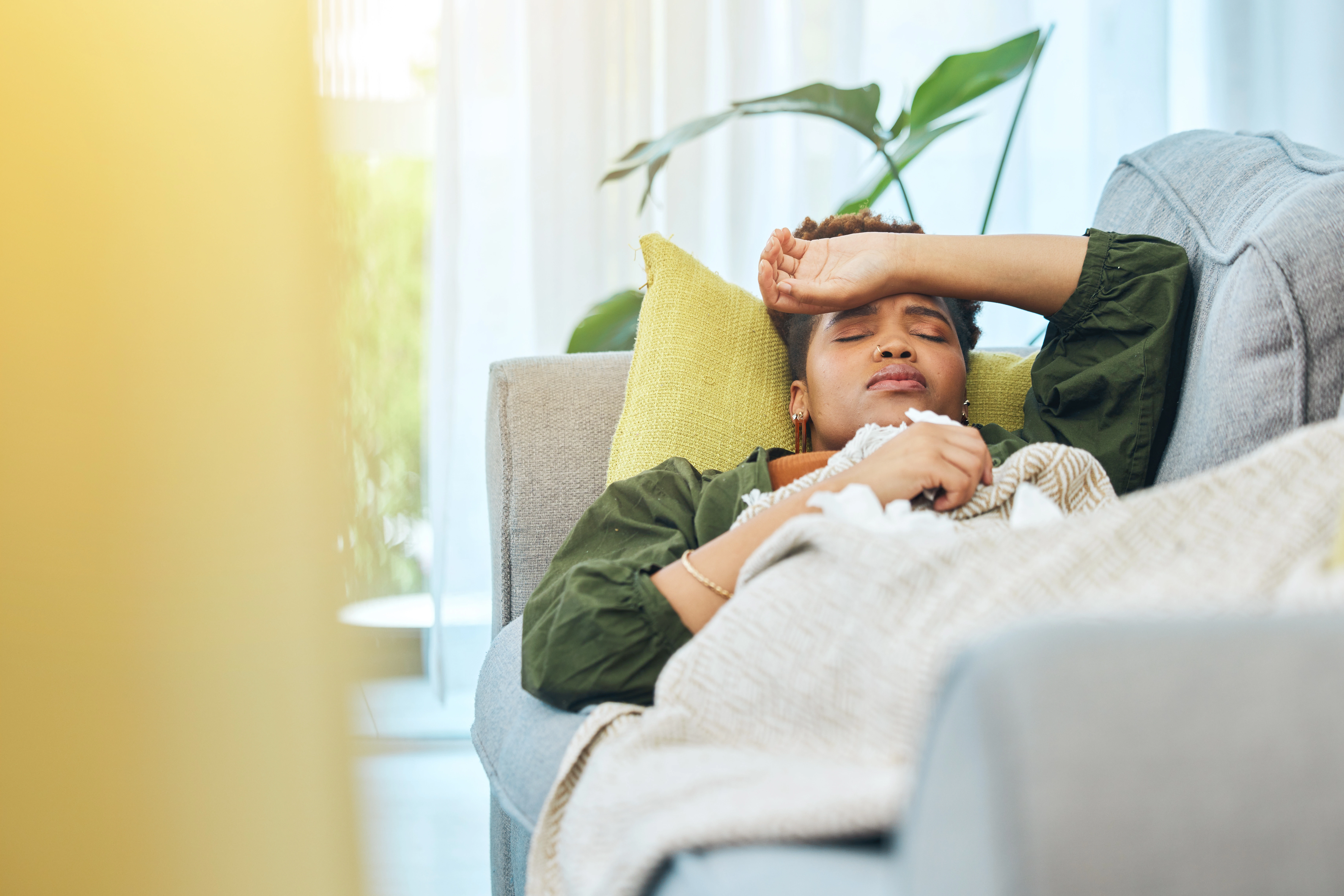 An adult woman with immune deficiency is laying on the couch with her hand to her forehead, indicating that she is unwell. 