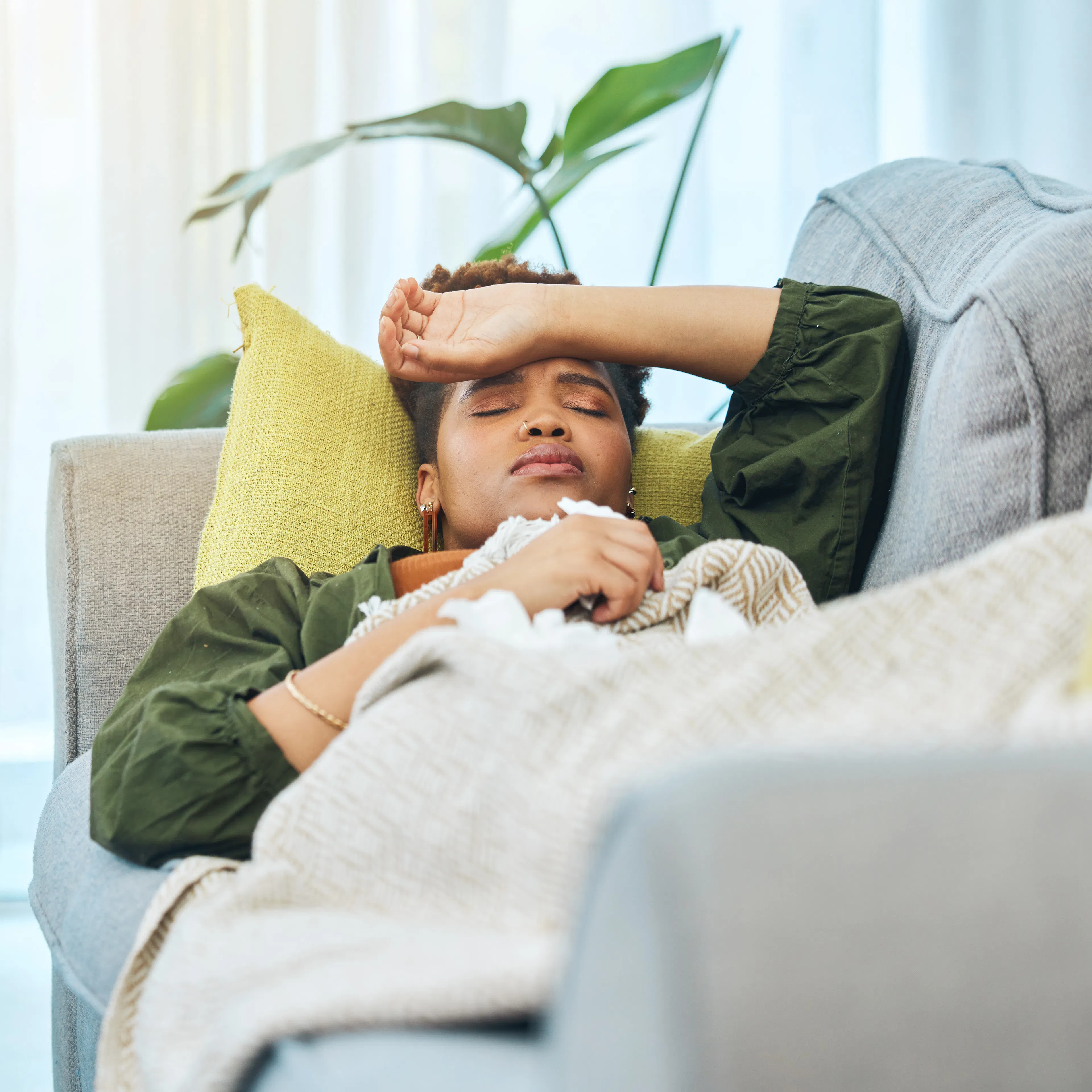 An adult woman with immune deficiency is laying on the couch with her hand to her forehead, indicating that she is unwell. 
