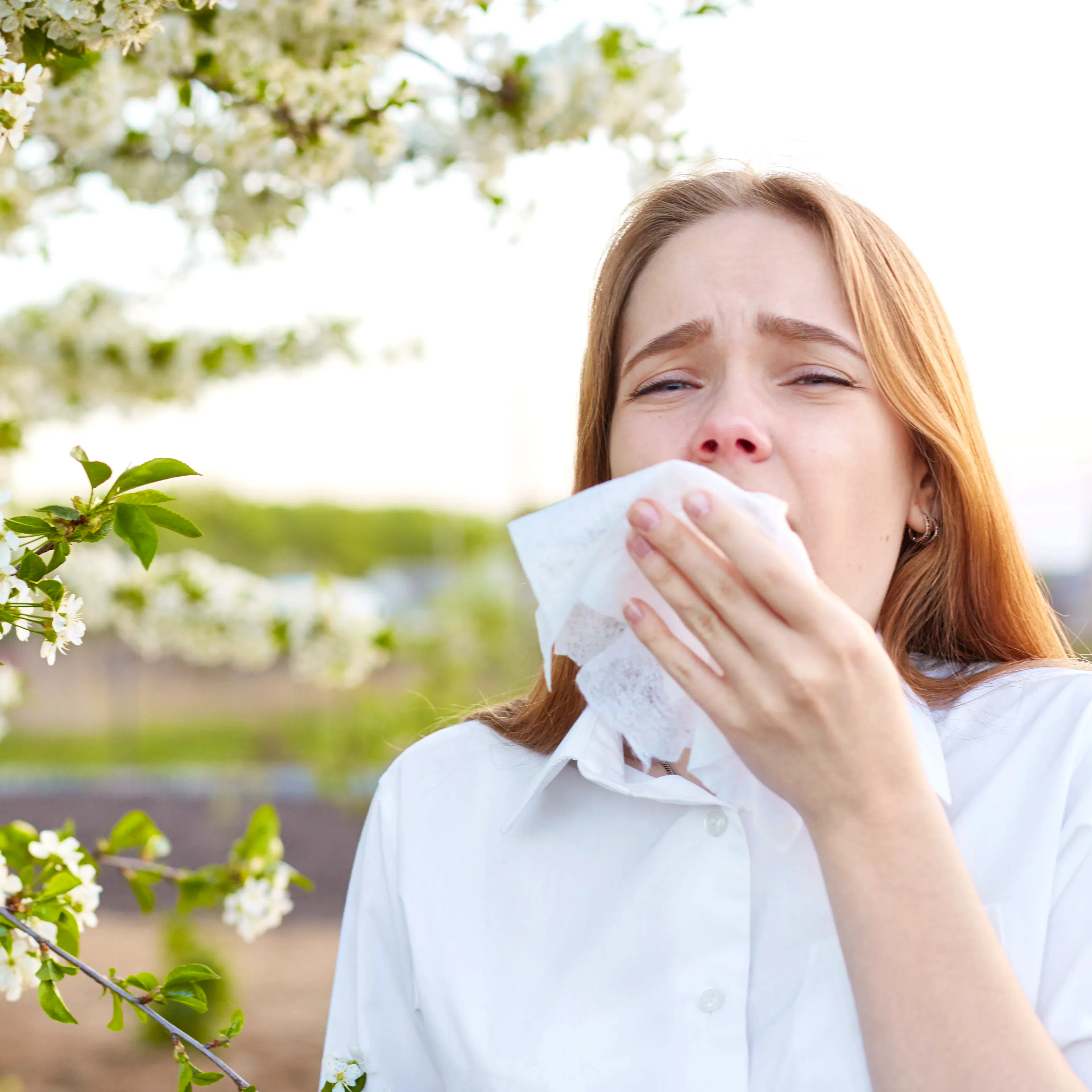 A woman with allergic rhinitis is blowing her nose, showing visible discomfort next to flowers.