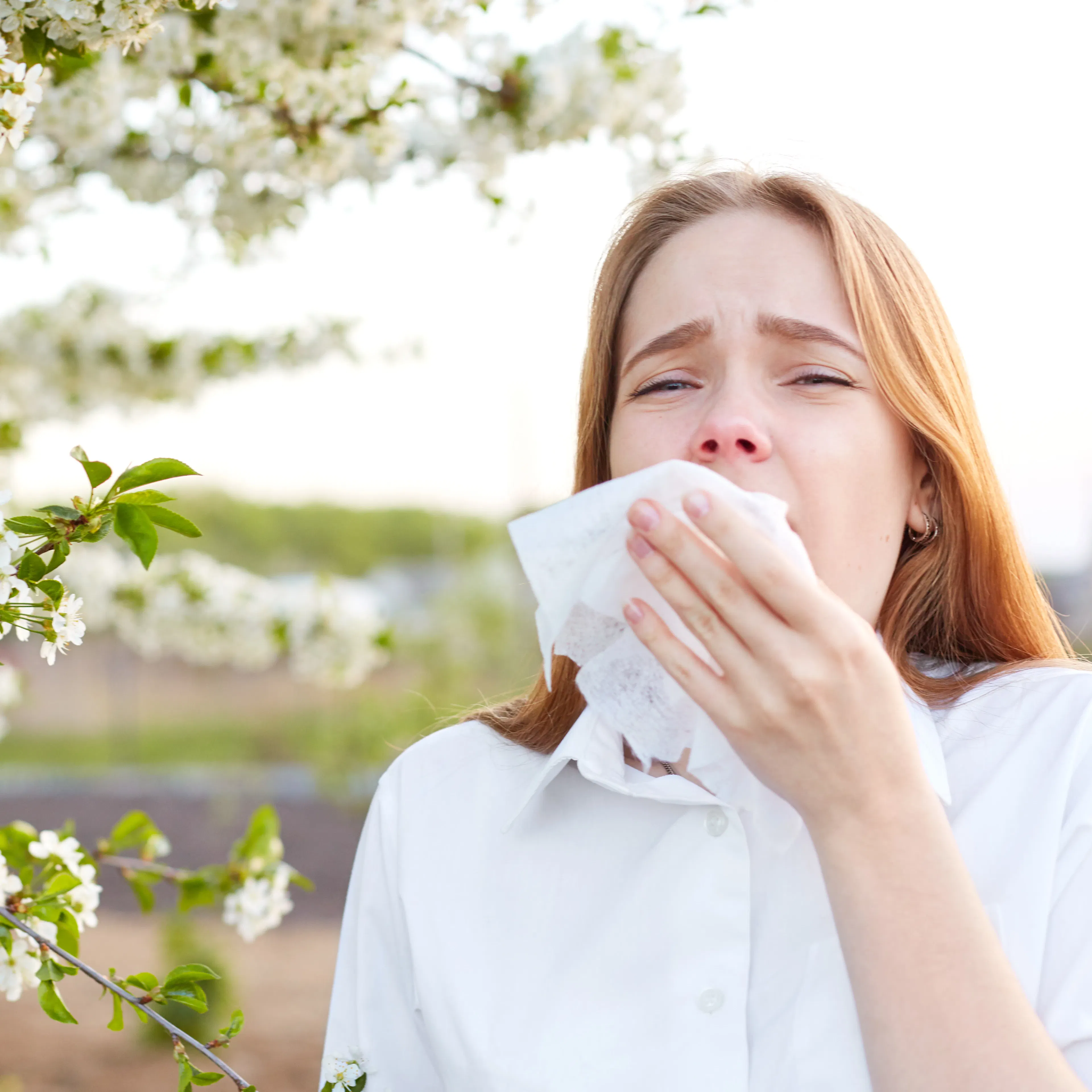 A woman with allergic rhinitis is blowing her nose, showing visible discomfort next to flowers.