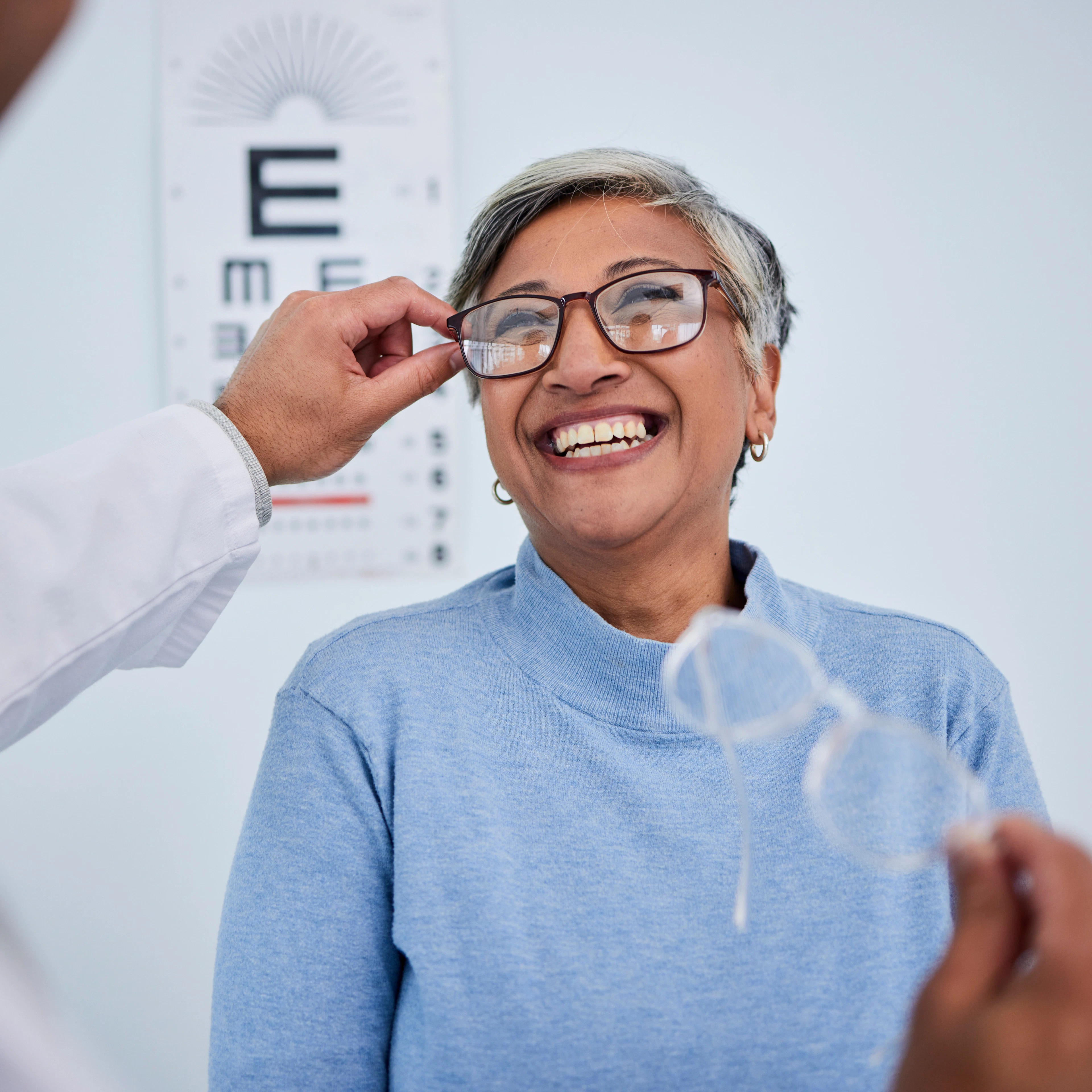 A middle-aged woman is trying on glasses happily at the eye care clinic with her optometrist. 