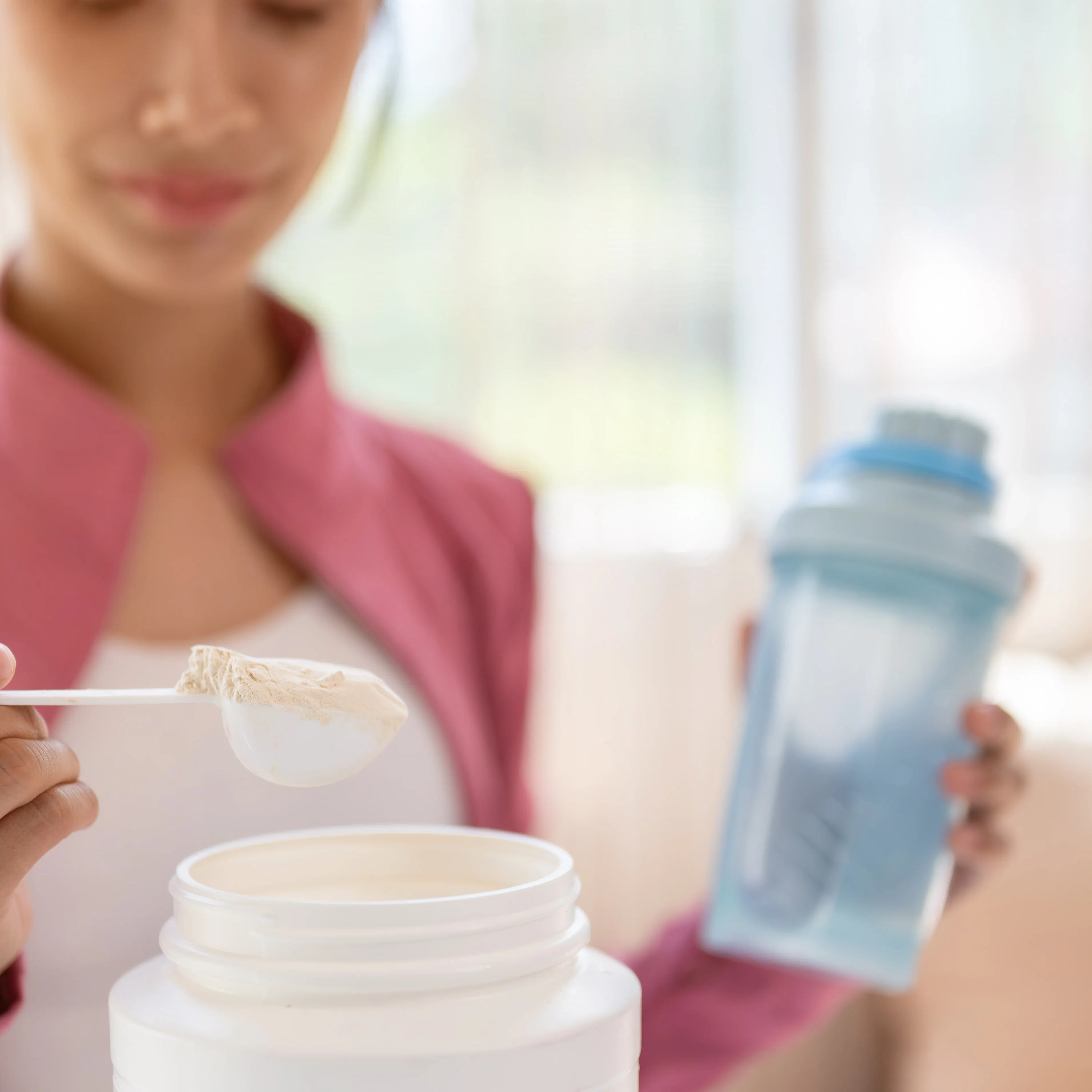 A woman in athletic wear is scooping a supplement, either creatine or protein powder, into her shaker bottle.