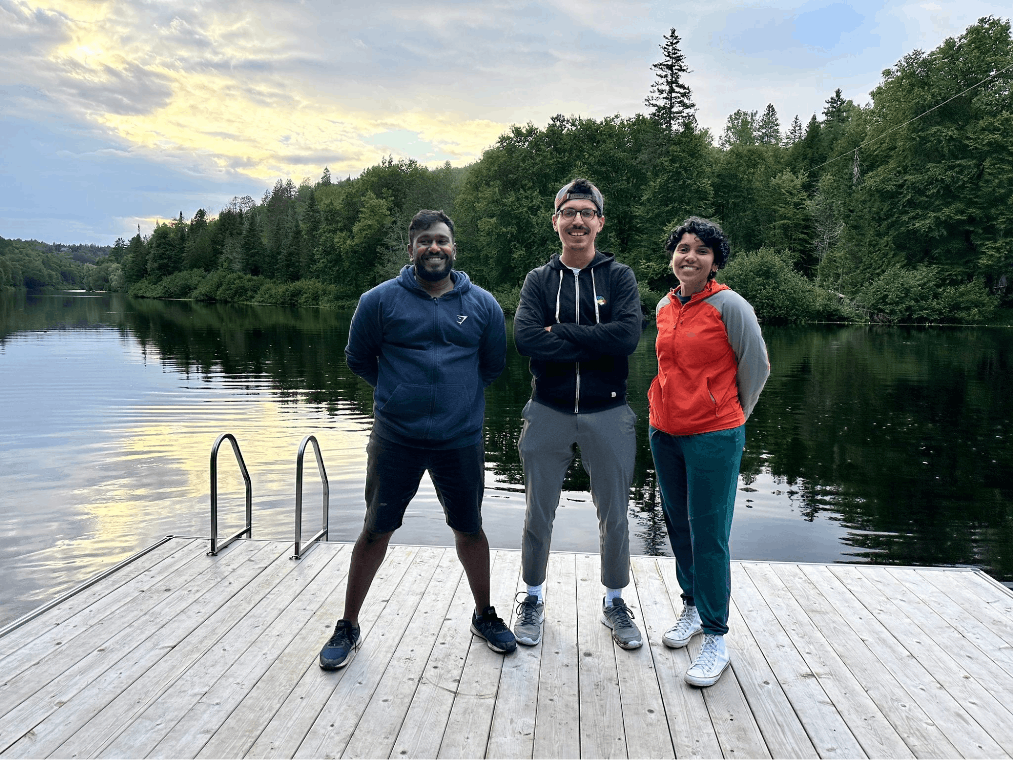 An image of Malik, Seby, and Sudnya on a dock. There are trees, water, and the Canadian skyline behind them.