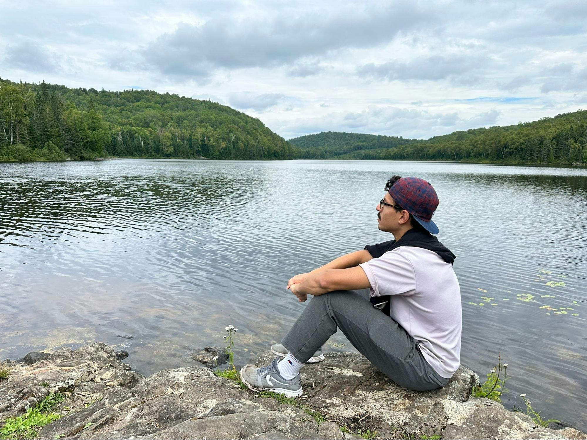 Malik outside on a hike. He is seated on the banks of a clear lake, with clouds and green mountains in the background.