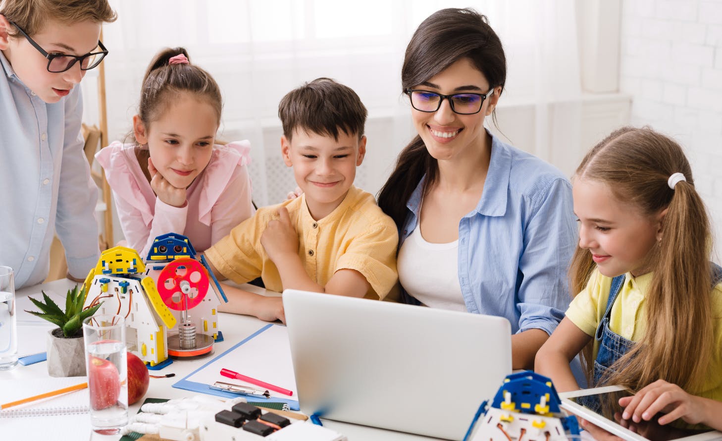 A group of students sit at a table with an adult. They look at a laptop together and there are pens, papers, books and toys on the table as well. 