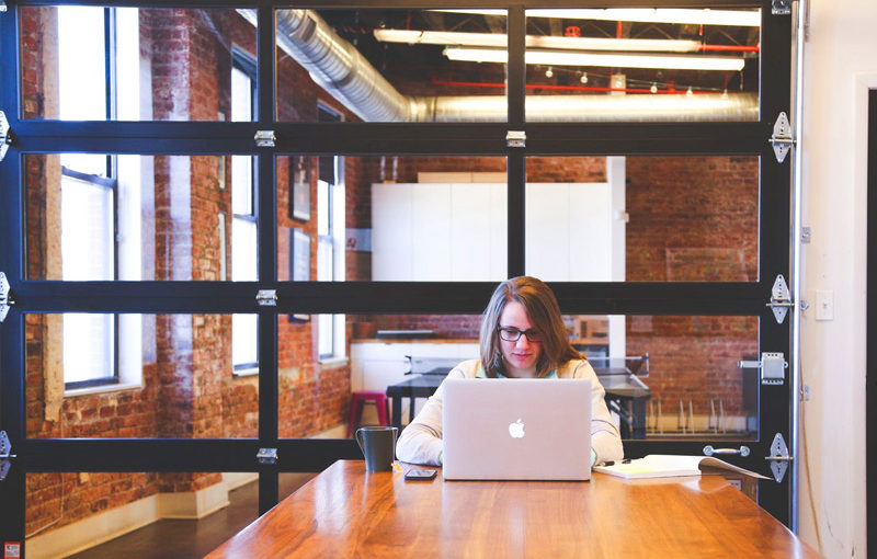 A woman works on a laptop at a wooden table in a modern, brick-walled office.