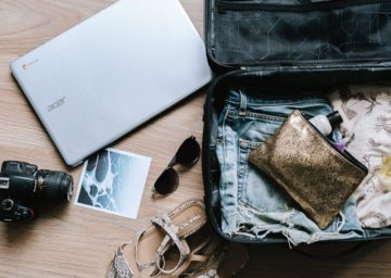 A suitcase packed with clothes, a laptop, sunglasses, a camera, and a gold wallet, all laid out on a wooden surface.