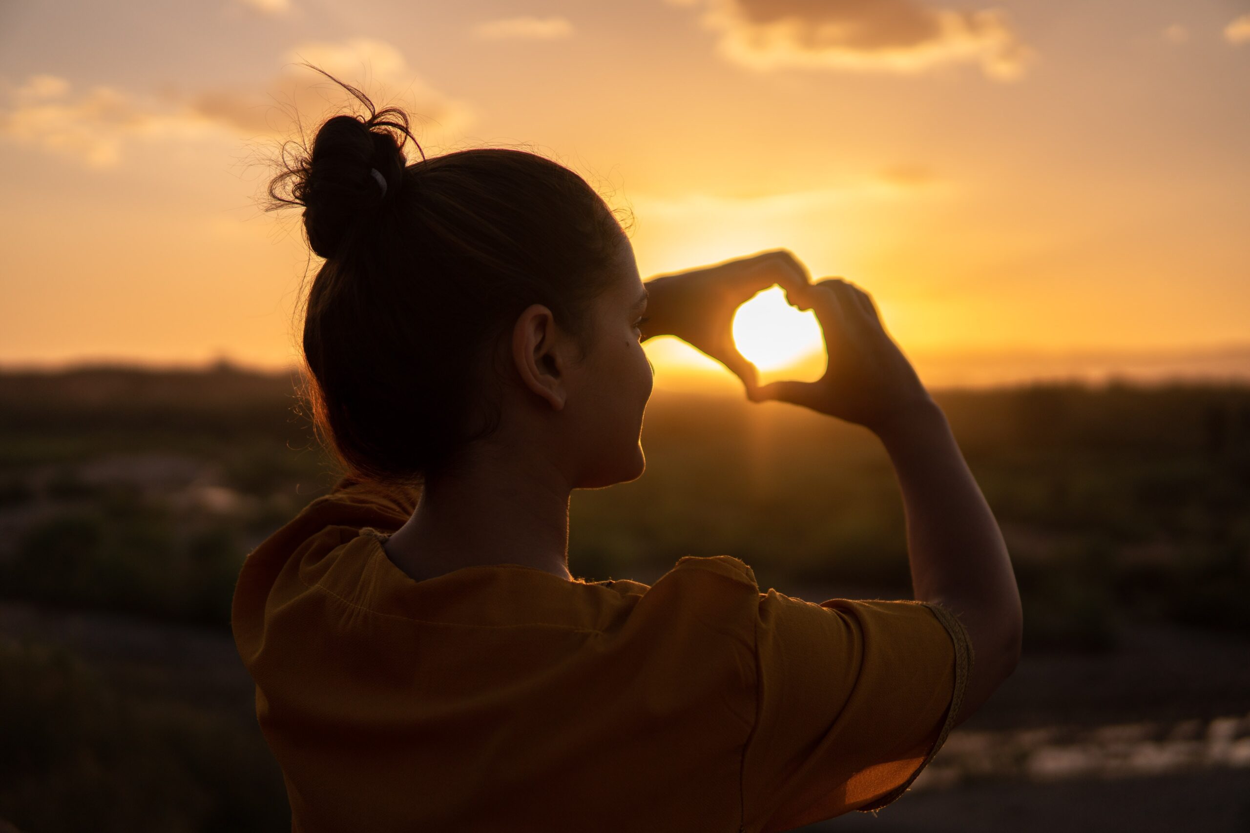 A woman at sunset forms a heart shape with her hands around the sun.