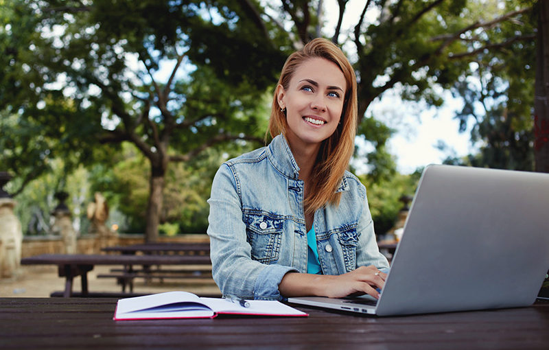 A smiling woman works on her laptop outdoors, surrounded by greenery and a notebook.