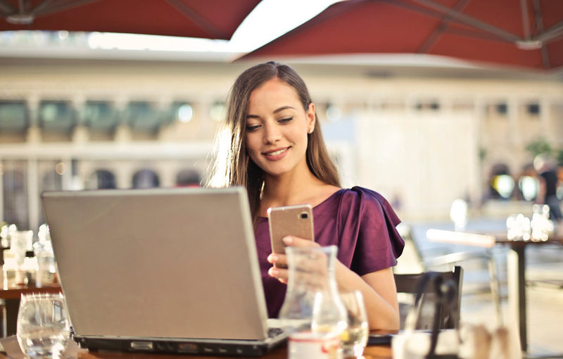 A woman smiles while using her phone at a café with a laptop nearby.