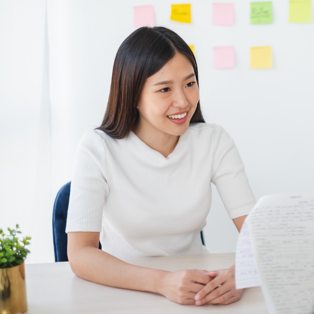 A young woman smiles while sitting at a table, with colorful sticky notes on the wall behind her, creating a cheerful atmosphere.