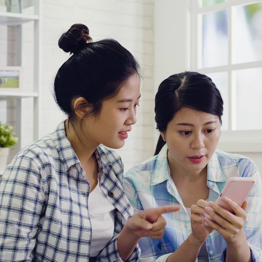 Two women sit together, one excitedly pointing at a smartphone.