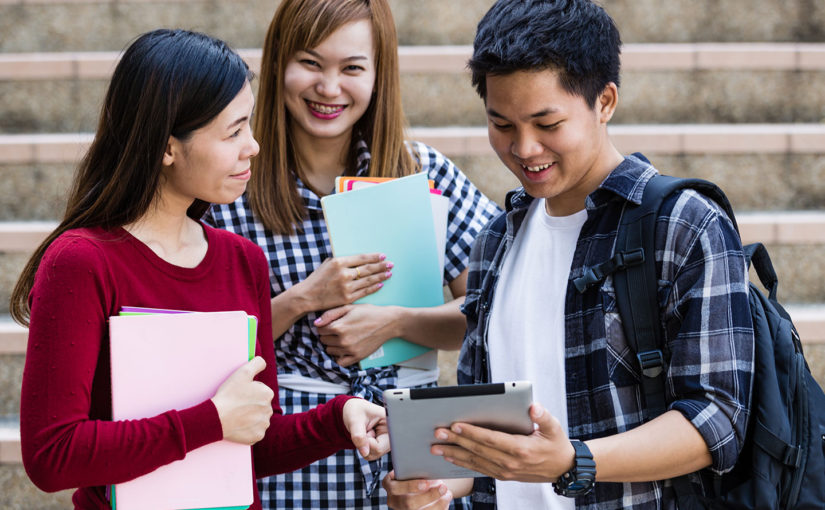 Three students outdoors, two holding folders and one with a tablet, engaged in discussion.