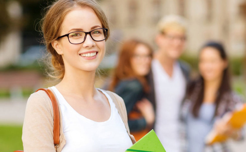 A smiling student with glasses holds books, surrounded by friends outdoors.