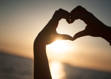 Silhouette of hands forming a heart shape against a sunset over the ocean.