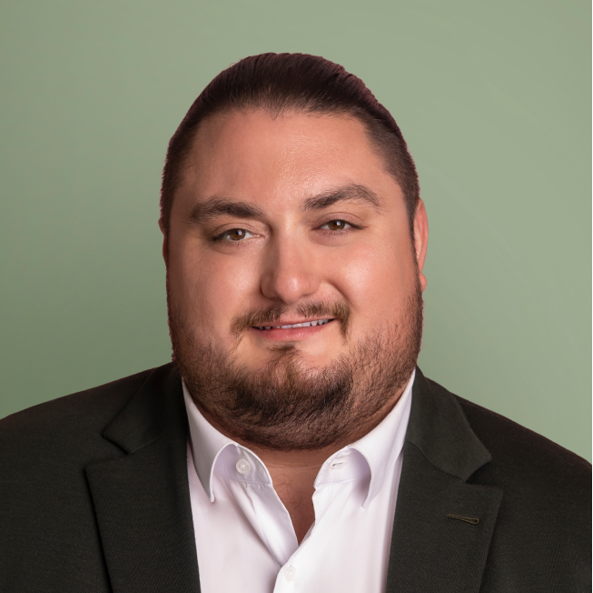 Portrait d'un homme souriant avec une barbe et les cheveux attachés en arrière, portant un costume foncé et une chemise blanche. L'arrière-plan est d'un vert doux.