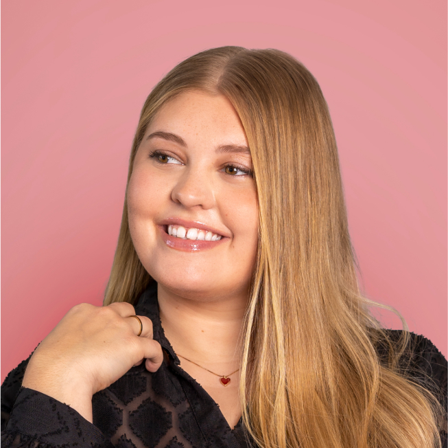 Portrait d'une femme souriante aux longs cheveux blonds, portant un haut noir texturé et un collier avec un pendentif en forme de cœur rouge. L'arrière-plan est d'un rose doux.