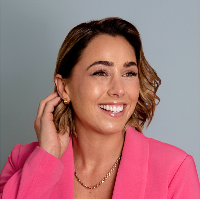 Portrait d'une femme souriante aux cheveux courts et ondulés, portant un blazer rose vif, un collier doré et des boucles d'oreilles assorties. L'arrière-plan est d'un bleu gris doux.