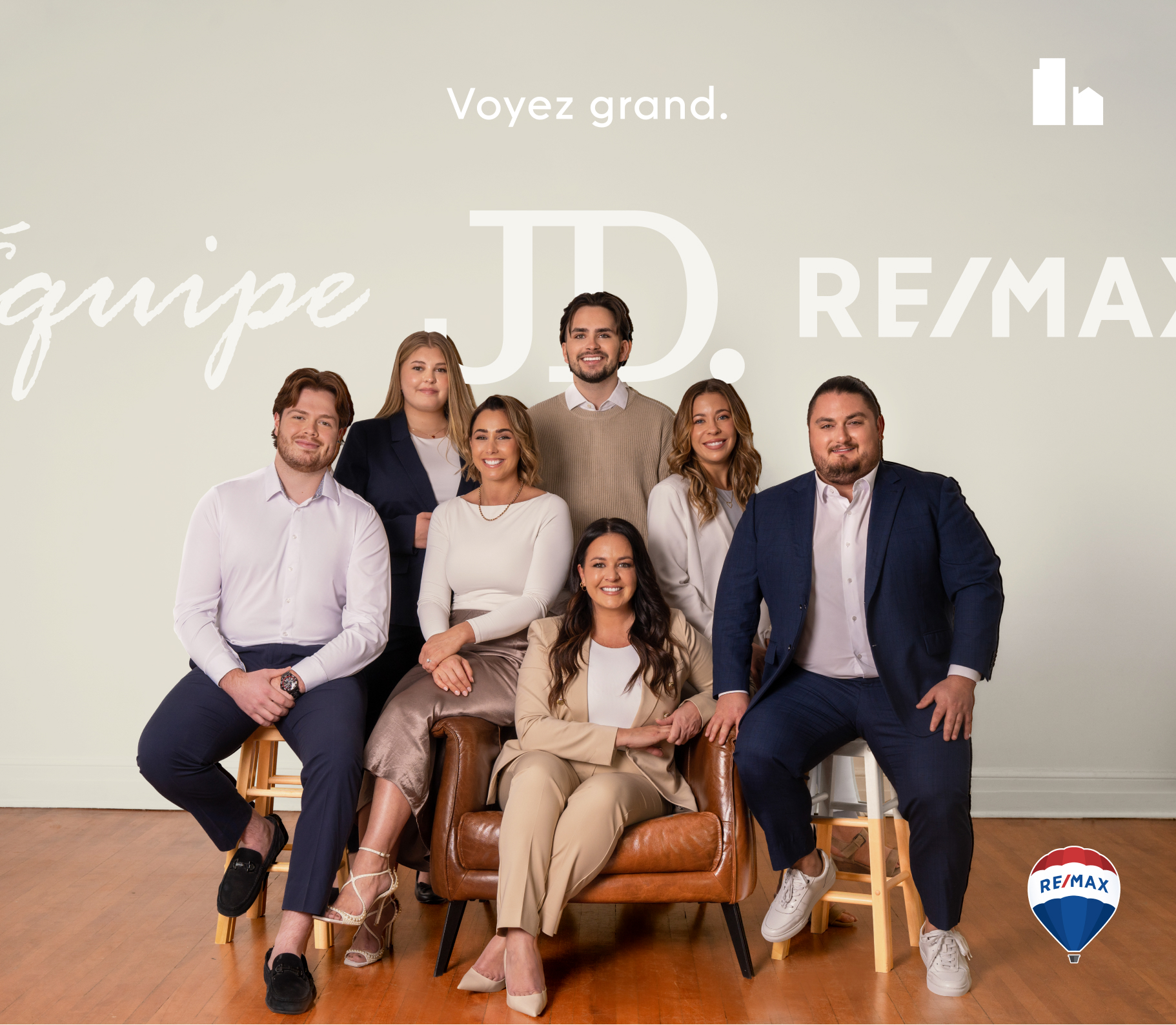 Groupe de sept professionnels posant dans un studio au décor minimaliste, avec le logo RE/MAX et le texte « Voyez grand. Équipe JD. » en arrière-plan.