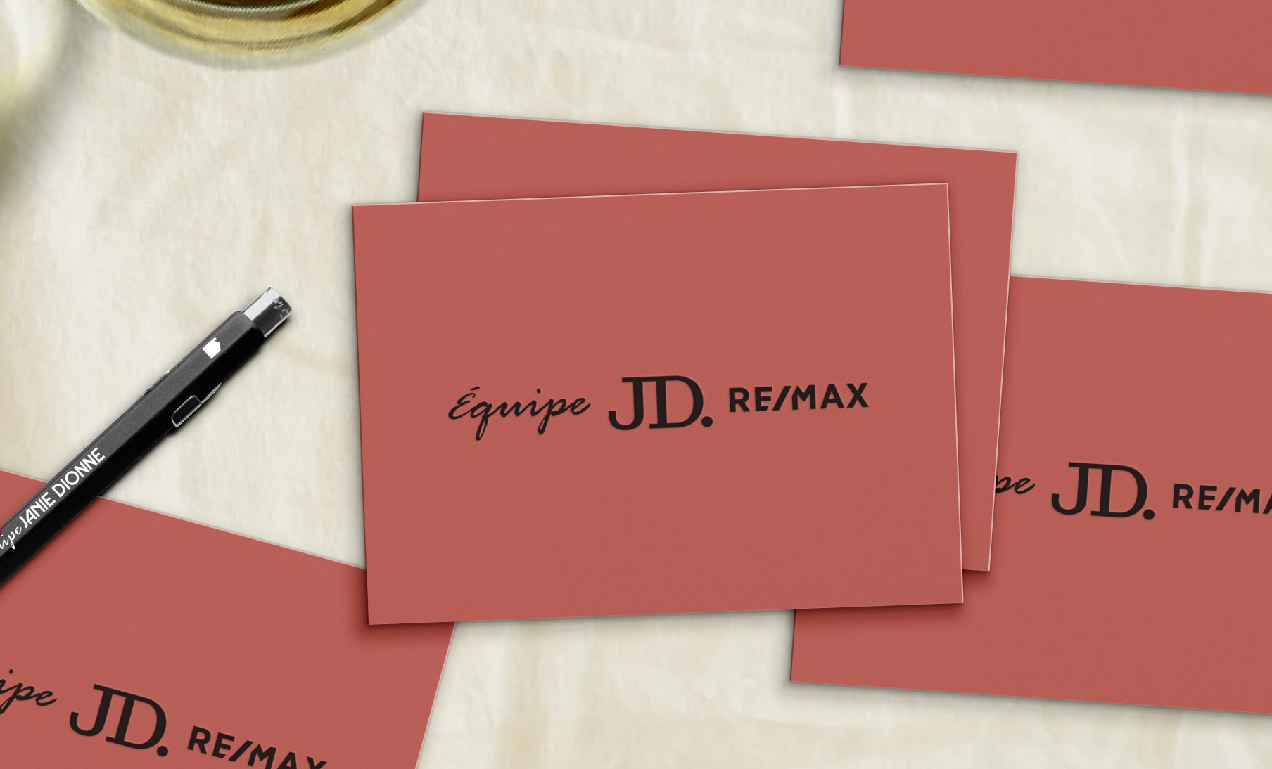 Cartes de visite terracotta avec l'inscription « Équipe JD. RE/MAX » en noir, posées sur une table avec un stylo noir marqué « Jannie Dionne ».